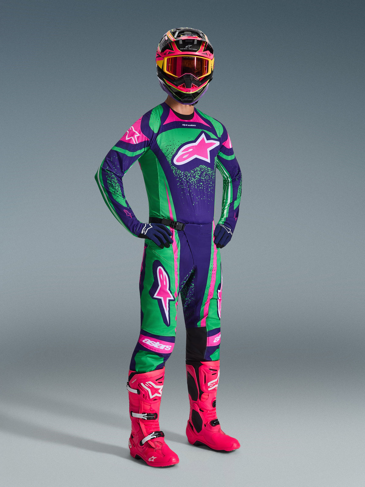 Motocross rider wearing the Alpinestars 2026 Techstar Nomur Gear Set in Deep Purple, Green, and Pink Fluo, featuring a matching jersey and pants, a colorful helmet with pink goggles, and vibrant pink boots, professional off-road racing equipment.