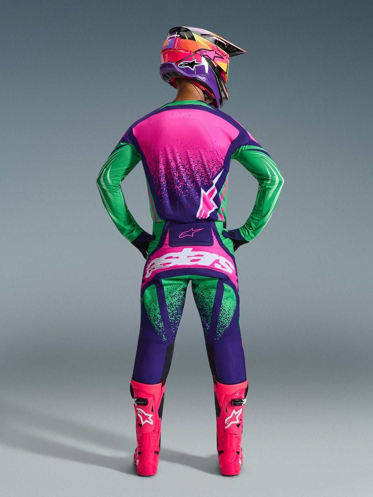 Alpinestars 2026 Techstar Nomur Gear Set, Motocross rider seen from behind wearing deep purple, green, and pink fluo jersey and pants, matching multi-colored helmet, bright pink fluo boots, coordinated professional racing outfit for off-road performance