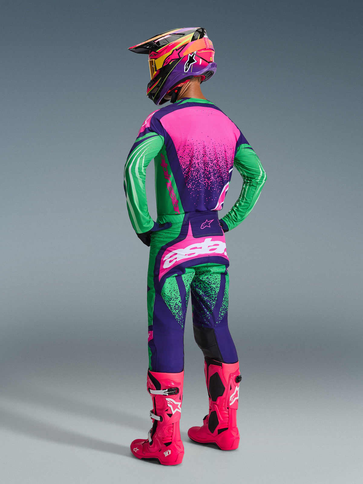Motocross rider wearing the 2026 Techstar Nomur Gear Set, Deep Purple Green and Pink Fluo, jersey with pink back and stippled texture, matching pants with white logos, bright pink fluo boots, and a colorful helmet, full-body back view for off-road racing performance