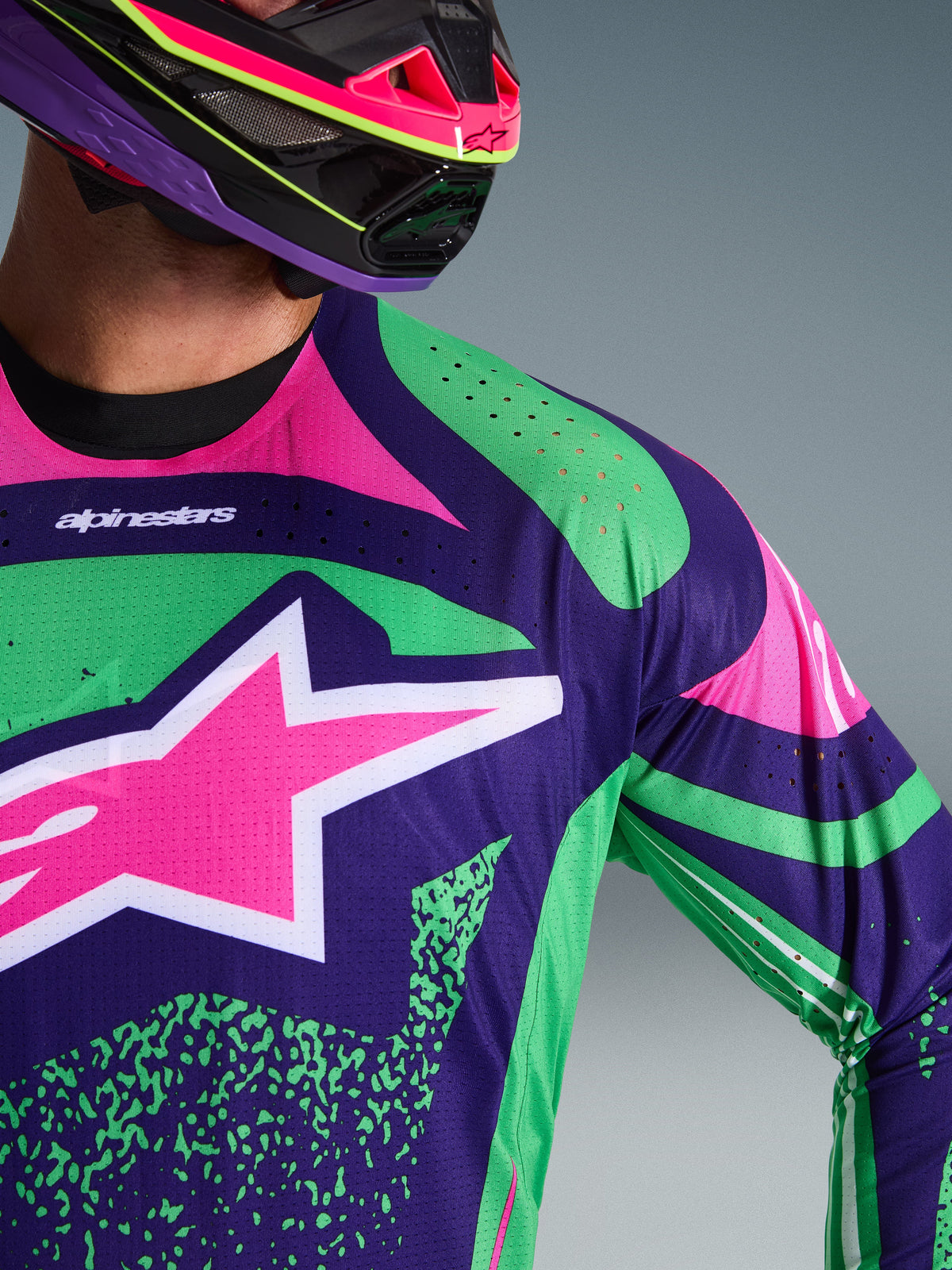 Close-up of Alpinestars 2026 Techstar Nomur Jersey, Deep Purple with Green and Pink Fluo accents, featuring a large pink star logo on the chest and laser-cut perforations on the shoulder, worn by a rider with a matching purple and pink motocross helmet.
