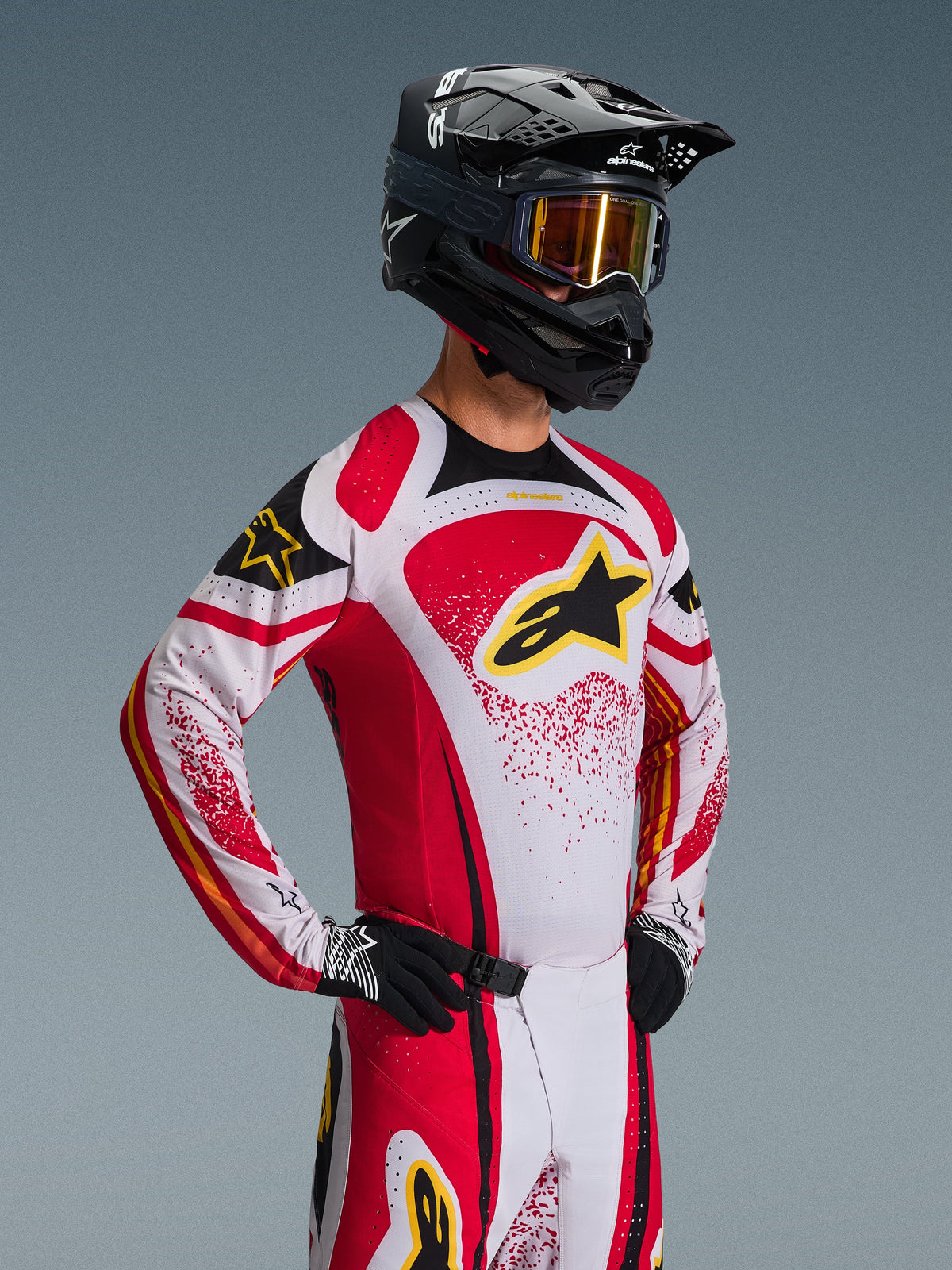 Motocross rider wearing the Alpinestars 2026 Techstar Nomur Gear Set, white and red jersey with black accents and yellow logo, matching pants, black helmet with gold-tinted goggles, black and white gloves, athletic race fit for off-road performance
