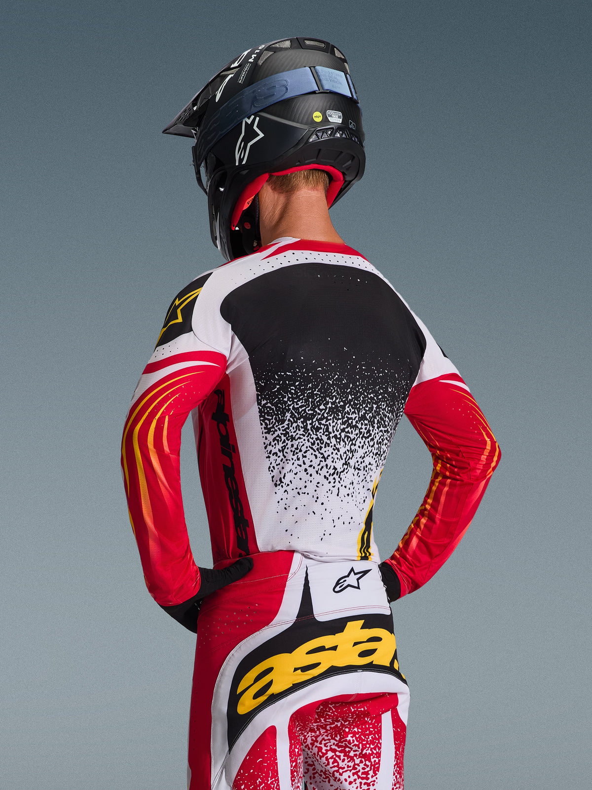 Alpinestars 2026 Techstar Nomur Gear Set, Motocross Jersey and Pants, Red White and Black with yellow accents, back view of rider wearing a black helmet, performance racing apparel with splatter graphics and bold branding