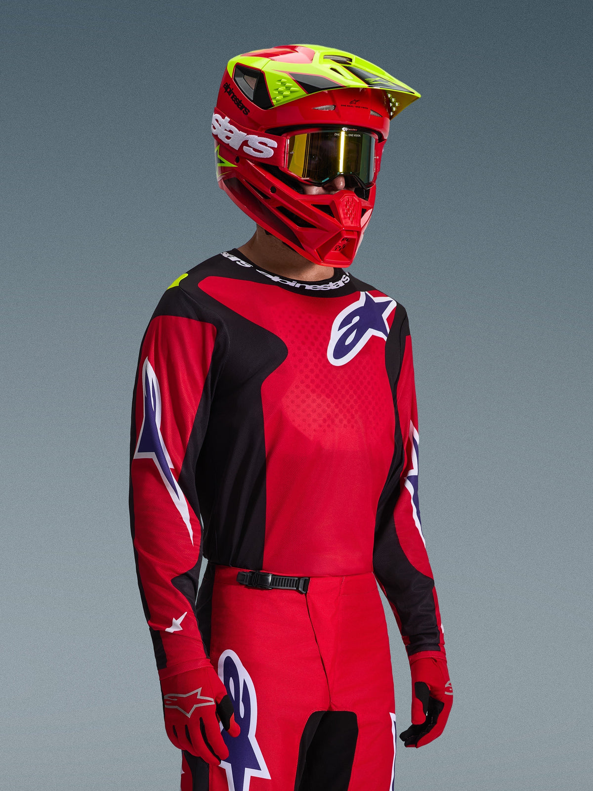 Motocross rider wearing Alpinestars 2026 Fluid Grid Gear Set, Red and Black jersey with white and blue logo accents, matching Red and Black pants, Red helmet with yellow highlights and gold goggles, coordinated off-road racing gear