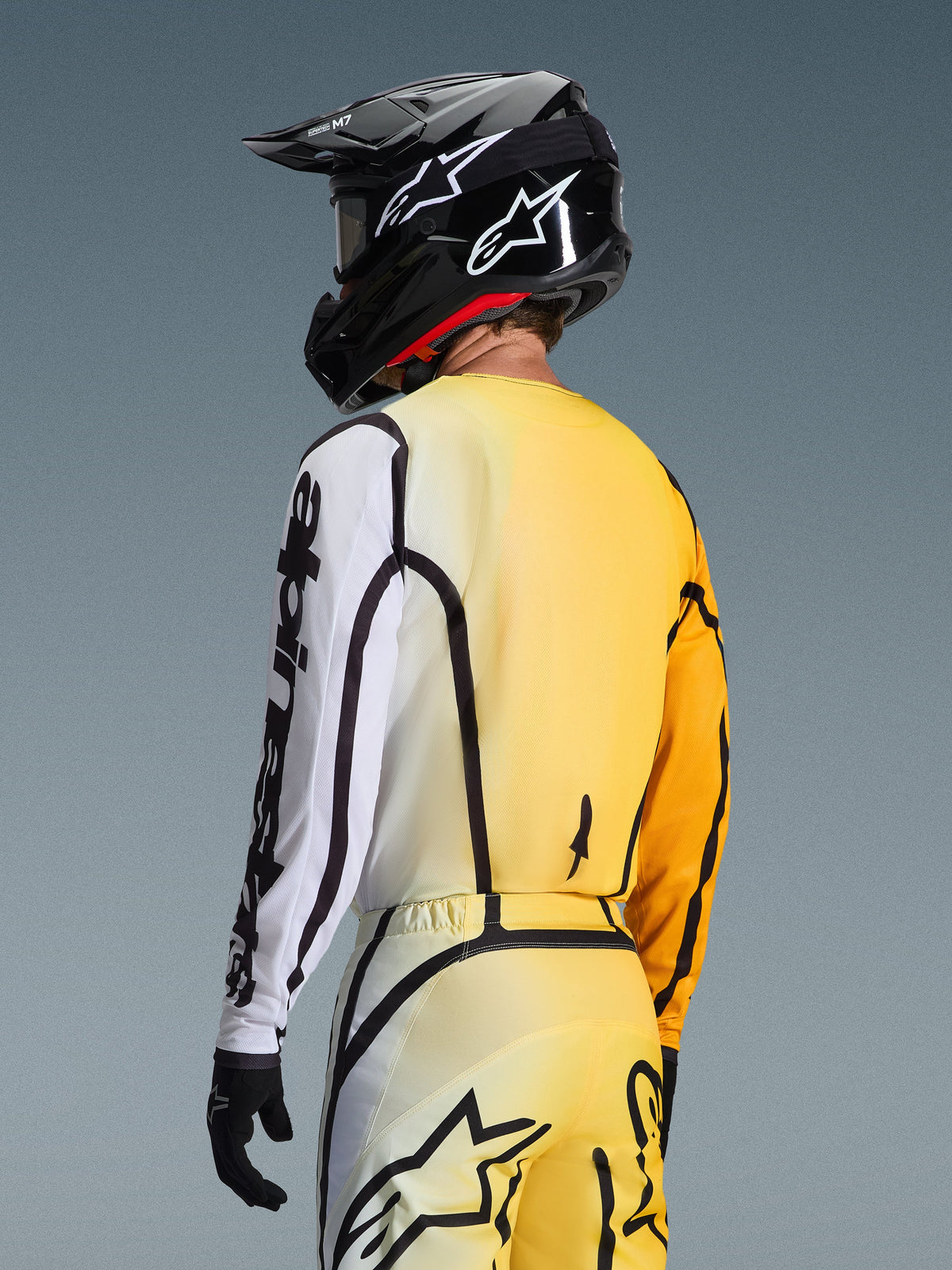 Rear three-quarter view of a motocross rider wearing the 2026 Fluid Apex Jersey in Yellow Black, featuring a gradient design from yellow to white, bold black Alpinestars branding on the sleeve, worn with matching pants and a black motocross helmet.