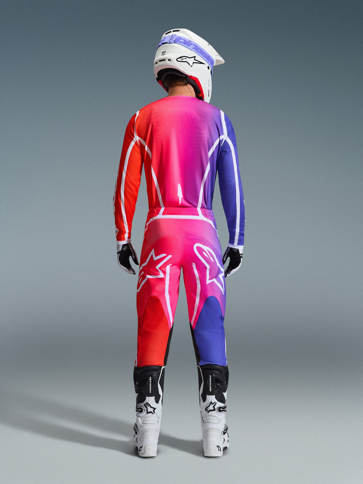 Alpinestars 2026 Fluid Apex Gear Set, Rear view of a motocross rider wearing a jersey and pants with a vibrant red, pink, and purple gradient, white graphic lines and large star logos, paired with a white helmet and white boots.