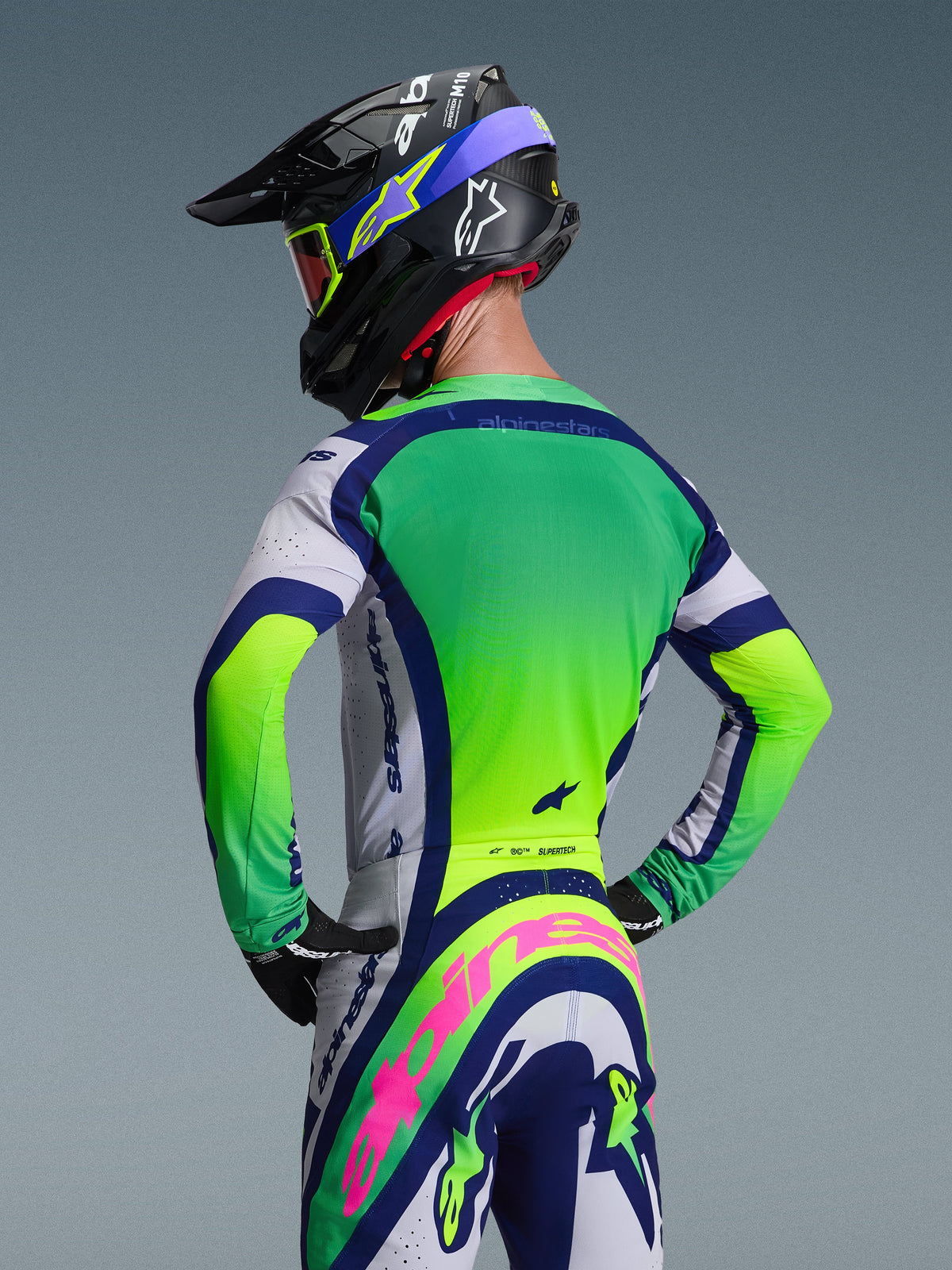 Rear view of a motocross rider wearing the Alpinestars 2026 Supertech Vista Gear Set, featuring a green and white jersey with navy blue accents, matching pants with a prominent pink Alpinestars logo, black racing helmet with blue graphics, and red-tinted goggles against a neutral gray background.