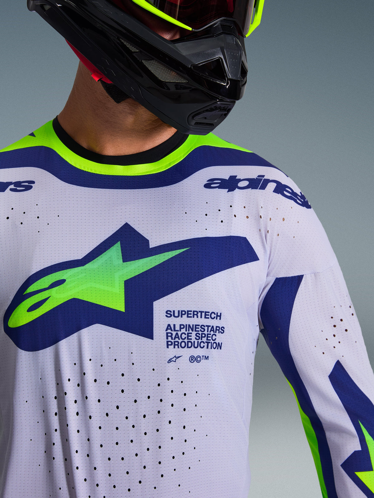 Close-up of the Alpinestars 2026 Supertech Vista Jersey in Light Gray, Yellow Fluo, and Blue, worn by a rider with a black helmet, featuring a large chest logo, laser-cut ventilation perforations, and professional race-spec branding.