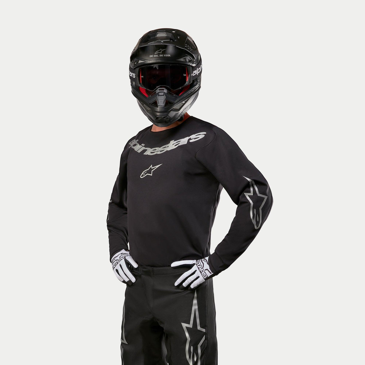 Motocross rider wearing Alpinestars 2024 Fluid Graphite Jersey, Black and Silver, paired with black motocross pants, black helmet with red-tinted goggles, and white and black gloves, featuring silver brand logos on the chest and sleeves.