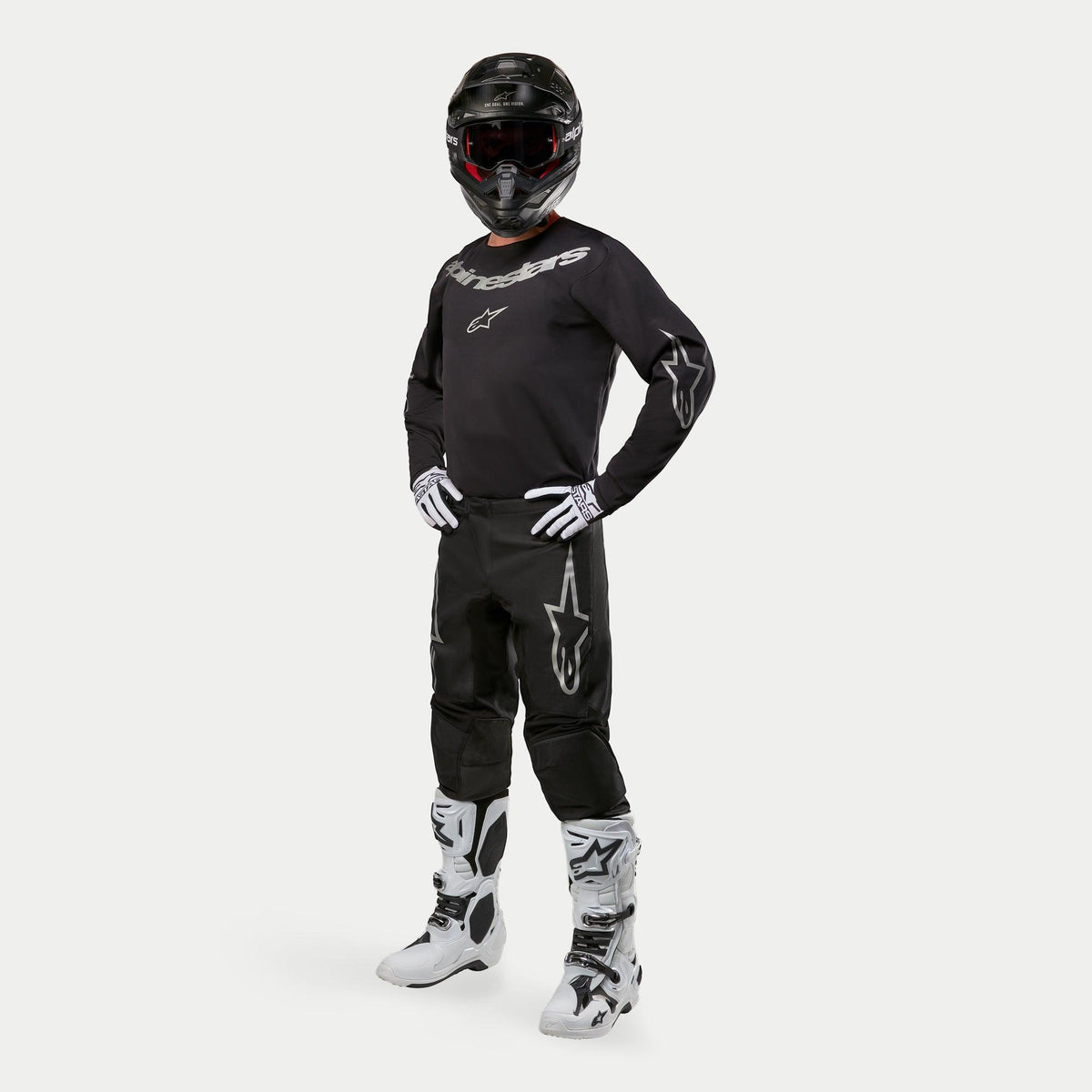 Motocross rider wearing Alpinestars 2024 Fluid Graphite Jersey in Black and Silver, matching black pants with silver star logos, white and black boots, white and black gloves, and a black helmet with red goggles, full off-road racing gear