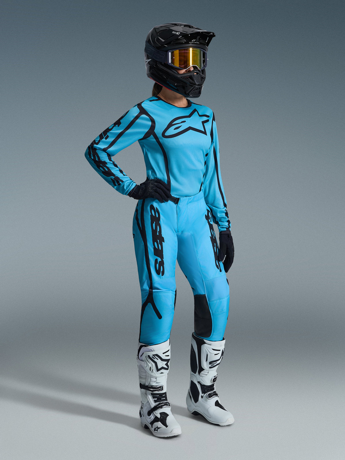 Alpinestars 2026 Woman Stella Fluid Apex Gear Set in Tropical Black, featuring a light blue jersey and matching pants with black accents, worn by a female rider with a black helmet, iridescent goggles, and white boots, professional motocross racing gear.