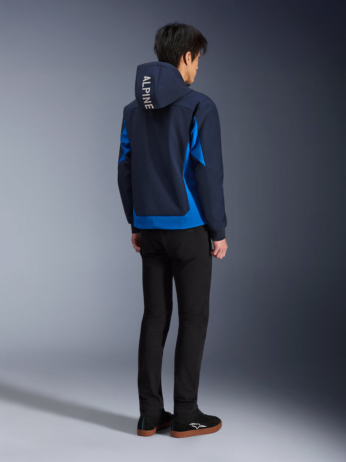 Alpinestars Radium Tech Hoodie, Night Navy and Royal Blue, back view of model wearing the technical shell hoodie with white 'ALPINE' text on the hood, paired with black pants and shoes, urban motorcycle riding gear