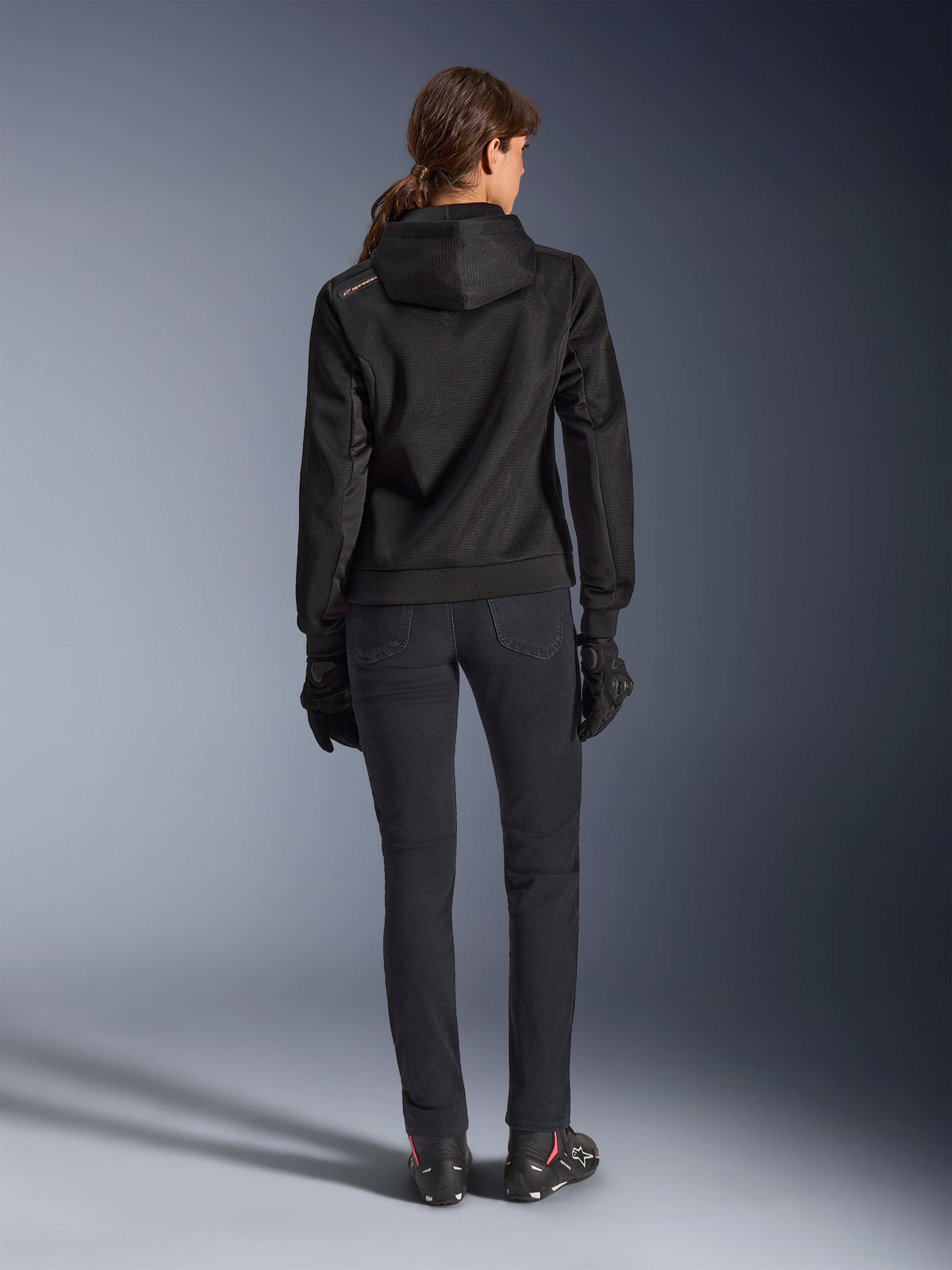 Alpinestars Stella Chrome Superairflow Hoodie, women's technical motorcycle hoodie, black 3D mesh with small orange logo on the shoulder, rear view, worn with black riding pants, black gloves and boots, designed for urban sport riding