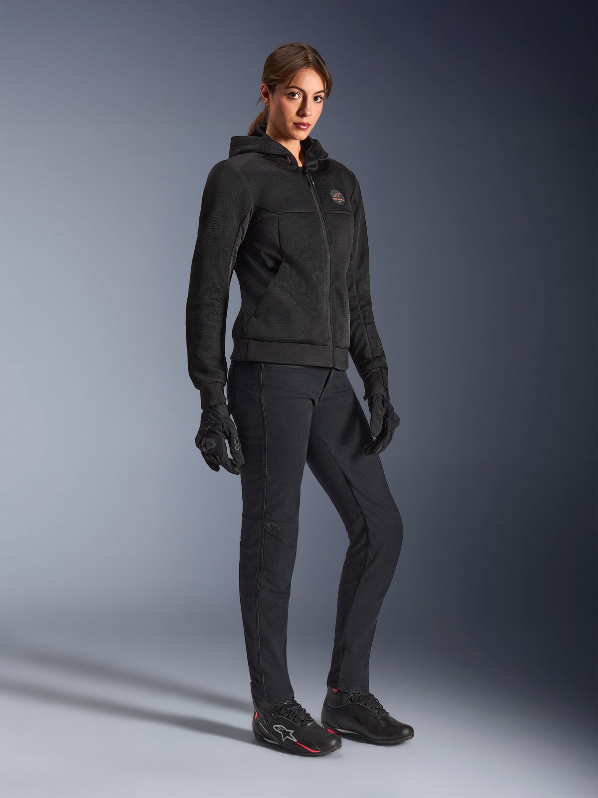 Female rider wearing the Alpinestars Stella Chrome Superairflow Hoodie in Black, technical motorcycle hoodie with 3D mesh construction, paired with black riding pants, gloves, and athletic-style riding shoes, urban sport aesthetic.