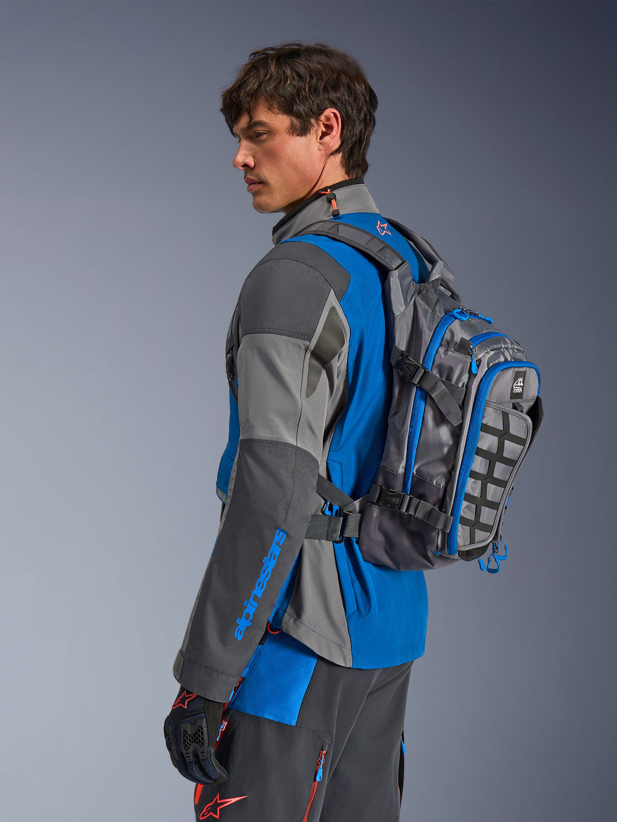 Alpinestars AT-15 Back Pack, Motorcycle Adventure Backpack, Dark Gray with Blue accents, worn by a rider in coordinated gray and blue technical riding gear, featuring MOLLE panel and rugged construction for off-road adventure.