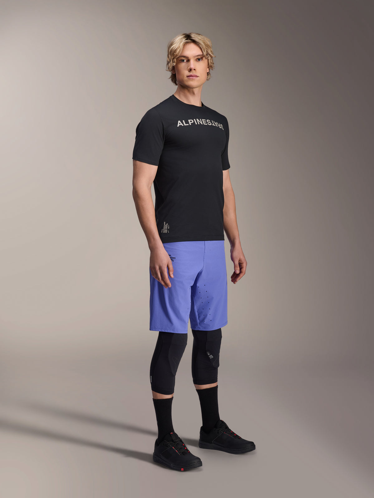 Male model wearing Alpinestars A-Aria Switch Short Sleeve Jersey in Black with white chest branding, paired with light blue mountain bike shorts, black knee guards, and black cycling shoes, studio full-body shot for performance MTB gear