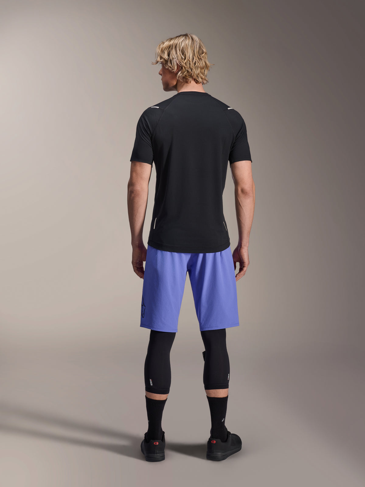 Alpinestars A-Aria Switch Short Sleeve Jersey, Black MTB Jersey, seen from the back, paired with blue mountain bike shorts and black leg protection, performance cycling gear for trail riding