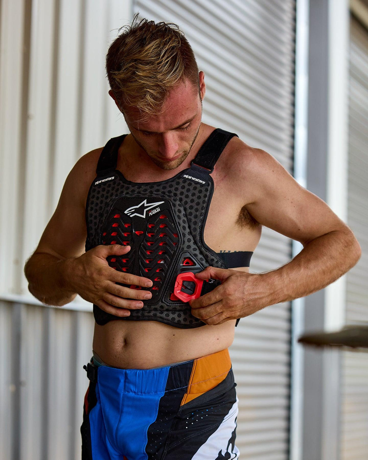 Man putting on the Alpinestars A-5 Plasma Chest Protector, Black with red accents and white logo, featuring a honeycomb mesh structure, worn with blue and orange motocross pants, preparatory setting for off-road racing