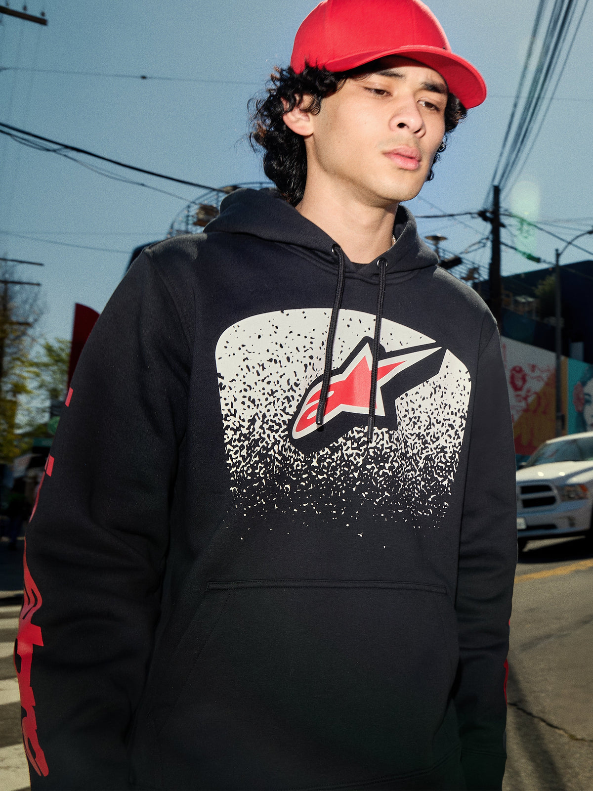 Alpinestars Obsessed Hoodie, black pullover fleece, white MX gradation chest graphic with red star logo, red wordmark logos on sleeves, casual motorsport-inspired streetwear worn in an urban environment.