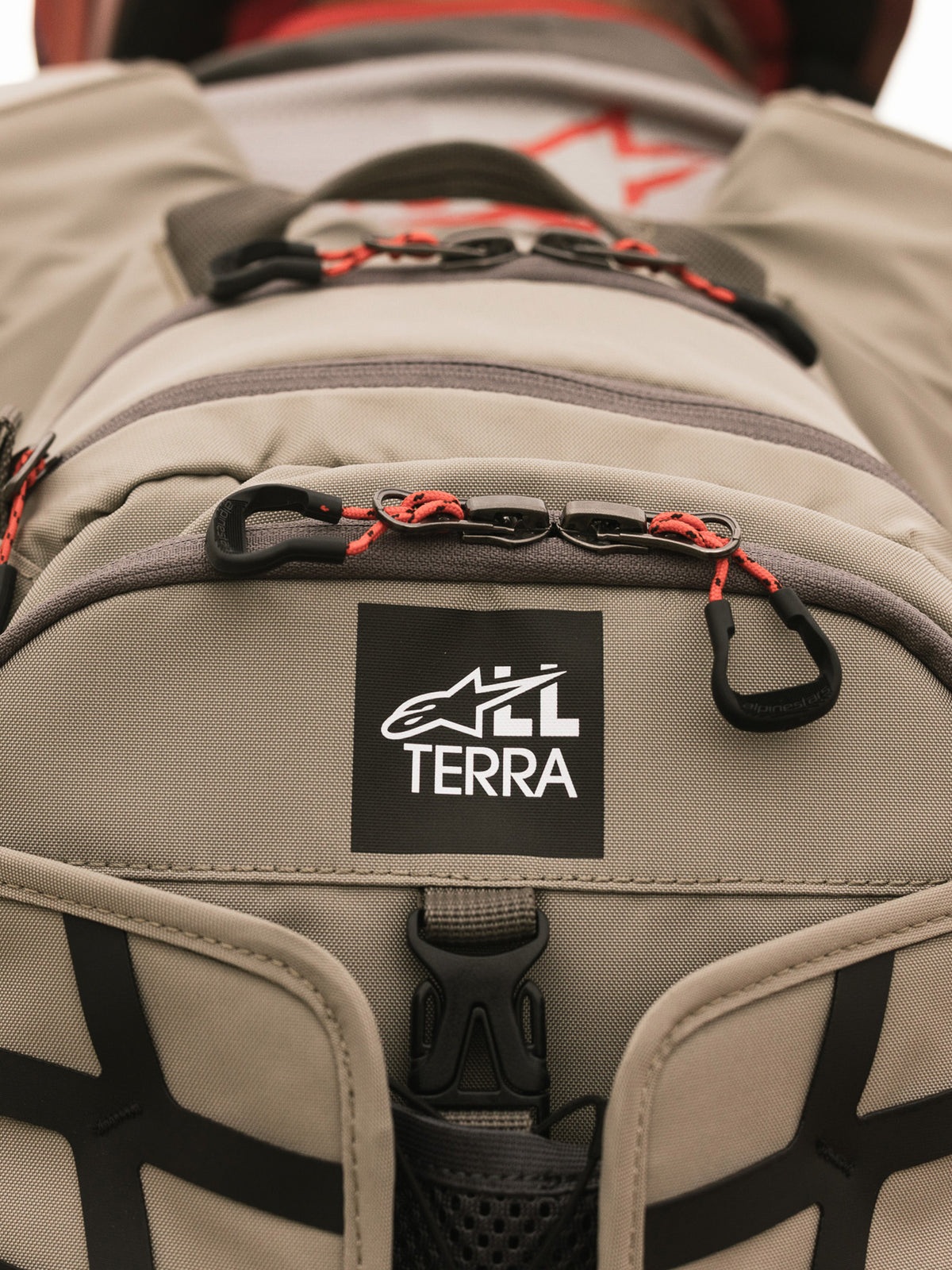 Alpinestars AT-15 Back Pack, Aluminum Walnut, close-up view of All Terra logo patch on tan technical textile, featuring orange corded zipper pulls and rugged construction details