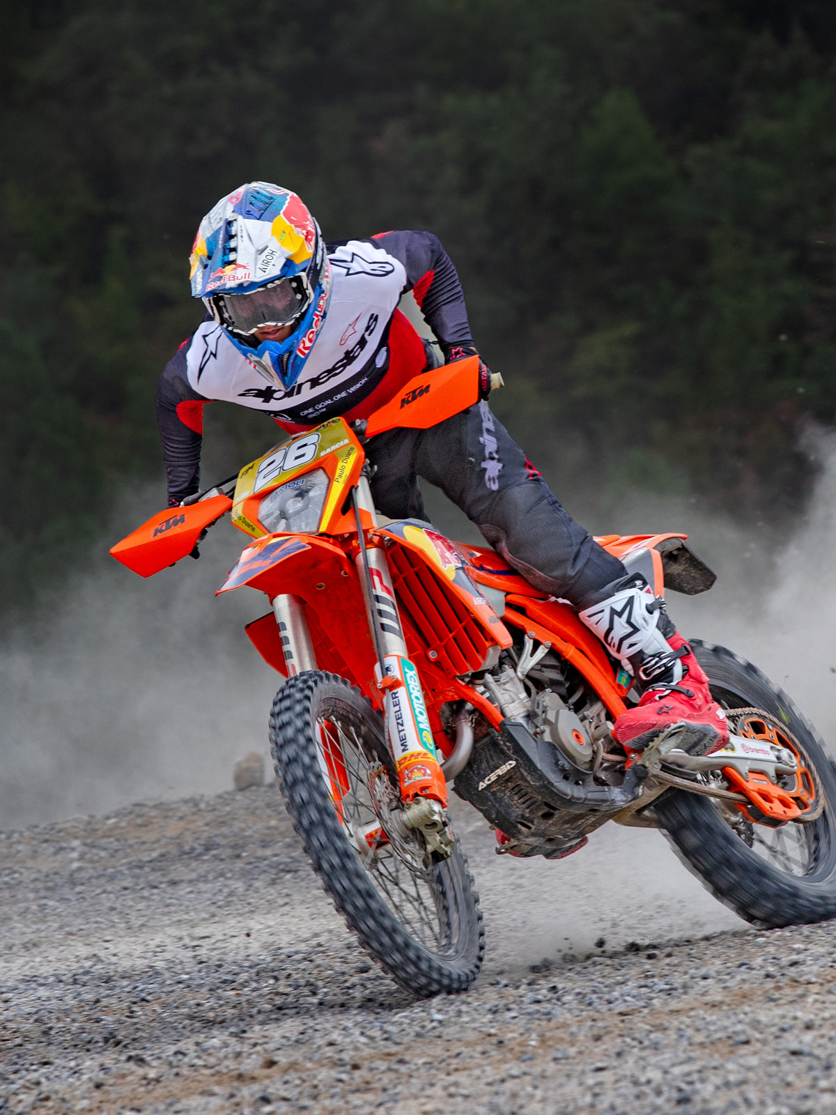 Motocross rider in action wearing Alpinestars Pro-Dura Jersey in Black, Bright Red, and White, riding an orange dirt bike on a dusty off-road trail, paired with black pants and red and white boots