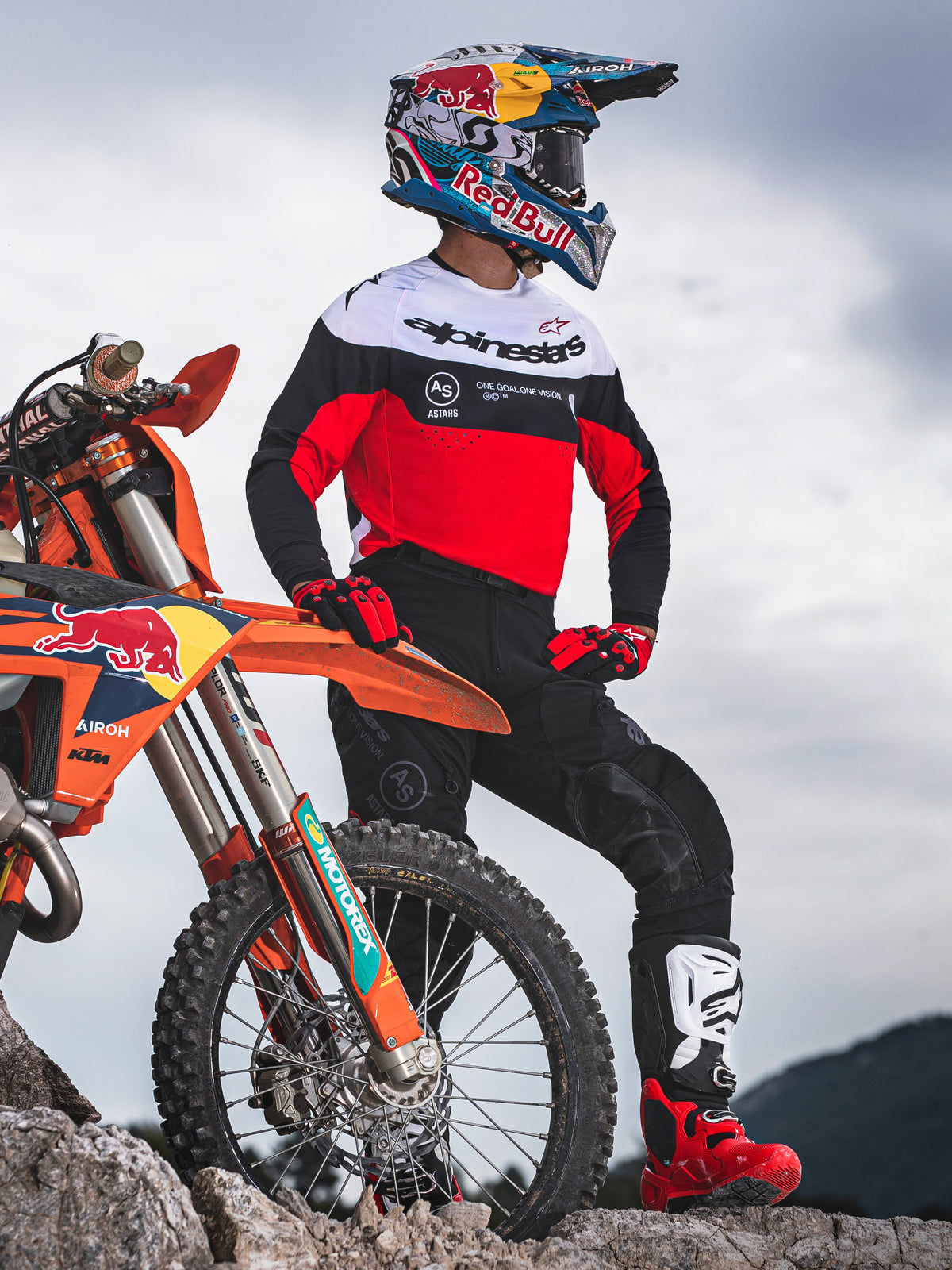Motocross rider standing beside an orange off-road motorcycle on rocky terrain, wearing Alpinestars Pro-Dura gear featuring a red, white, and black jersey and black pants, accessorized with red and black gloves, white and red boots, and a Red Bull helmet, complete professional enduro outfit.