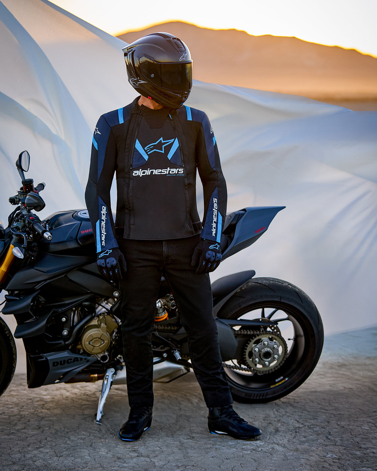 Alpinestars T-Stunt Air Jacket, Sport-Cut Textile Jacket, Black Dark Blue and Bright Blue, worn by a rider standing next to a Ducati motorcycle, featuring a carbon fiber helmet and black riding gear, set against a desert landscape at sunset.