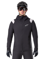 Alpinestars A-Dura Rain Jacket, MTB Rain Jacket, Black with white shoulder panels, worn with black MTB helmet, sunglasses, and gloves, waterproof design with hood and front zipper, sporty outdoor gear for cycling
