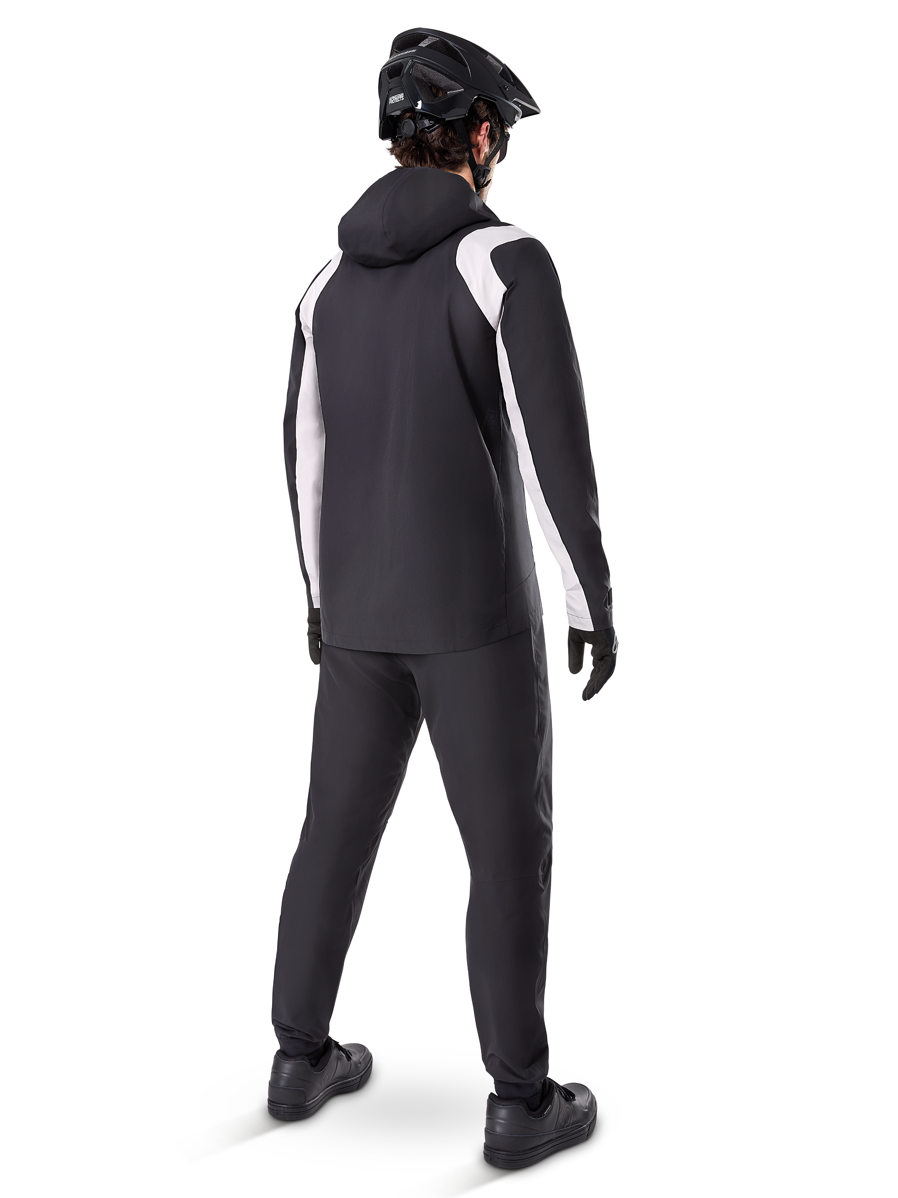 Alpinestars A-Dura Rain Jacket, back view of a male cyclist wearing the black and white waterproof MTB jacket, matching black cycling pants, black helmet, and gloves, technical gear for mountain biking in wet conditions.