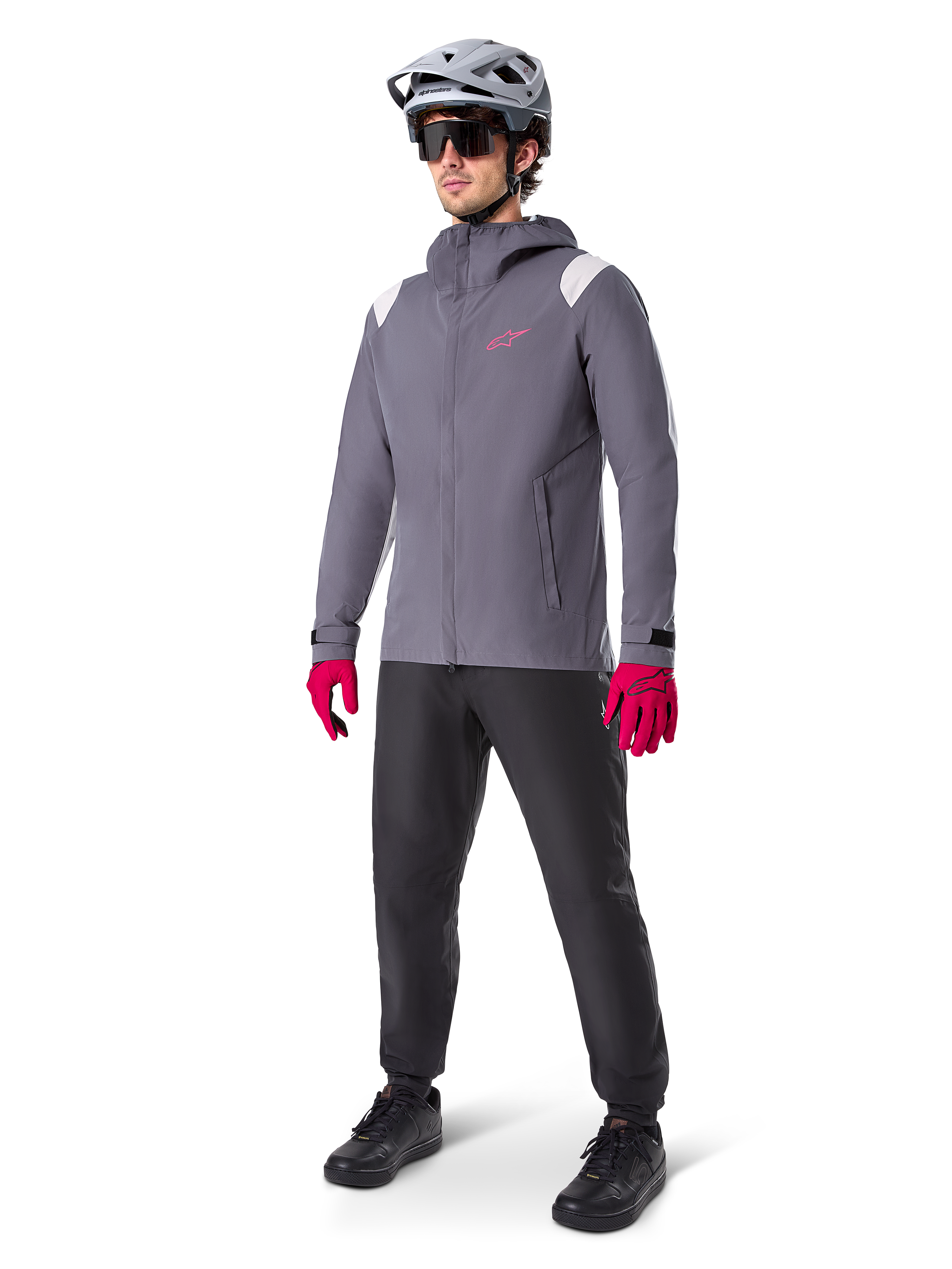 Male model wearing Alpinestars A-Dura Rain Jacket in Dark Titanium, gray hooded jacket with pink chest logo and light gray shoulder panels, paired with a gray MTB helmet, dark sunglasses, vibrant pink gloves, and black pants for off-road cycling.