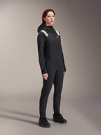 Woman wearing the Alpinestars Stella A-Dura Rain Jacket in Black, 2.5-layer waterproof MTB shell with light grey shoulder accents, matching black pants and cycling shoes, studio portrait for performance cycling gear