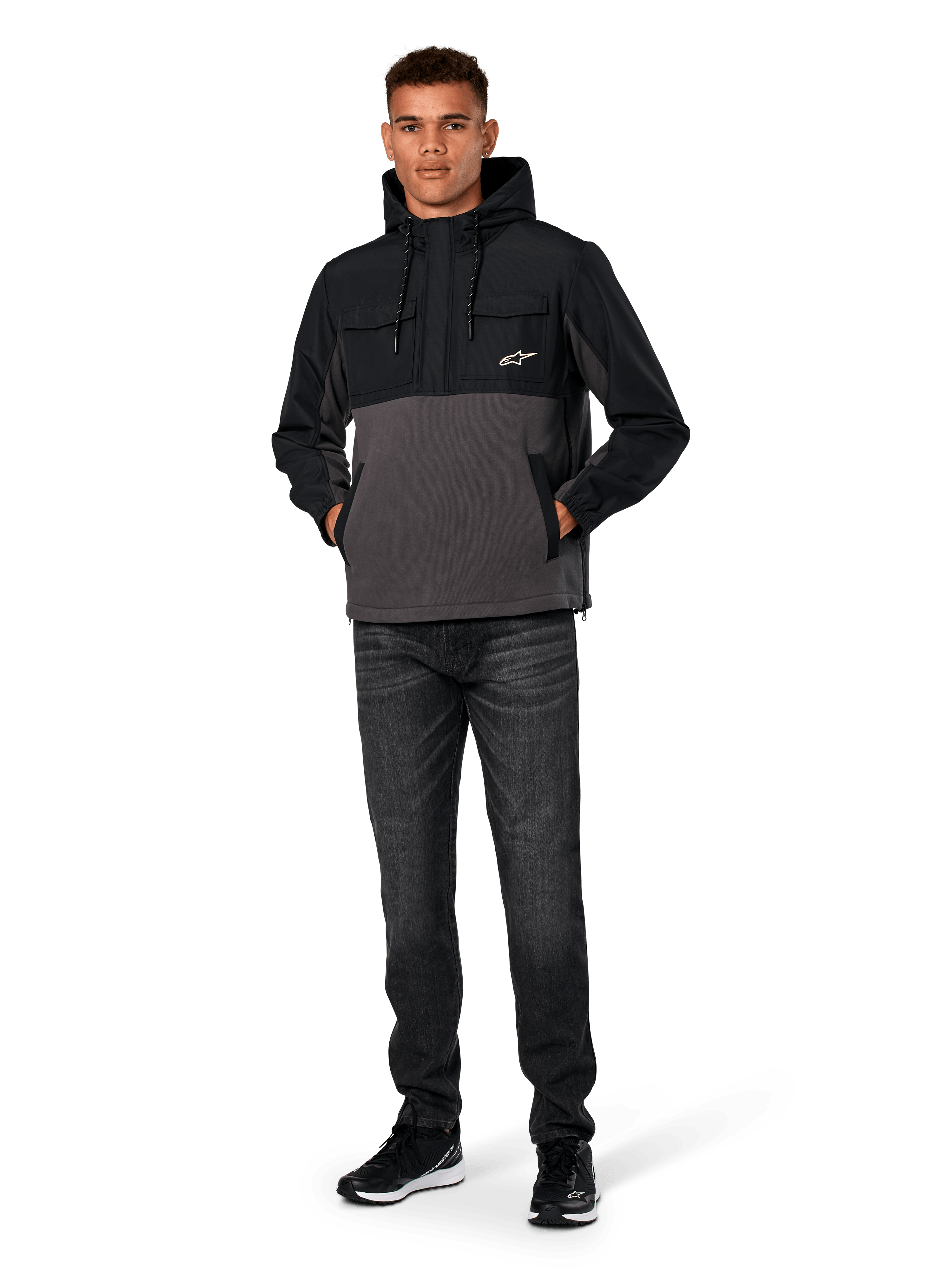 Alpinestars Juncture Hybrid Jacket, Black and Gunmetal, mixed-material anorak hoodie featuring a canvas fleece body and ripstop overlays, white A-star logo on chest, drawstring hood, worn by model with dark jeans and black sneakers, casual motorsport-inspired streetwear