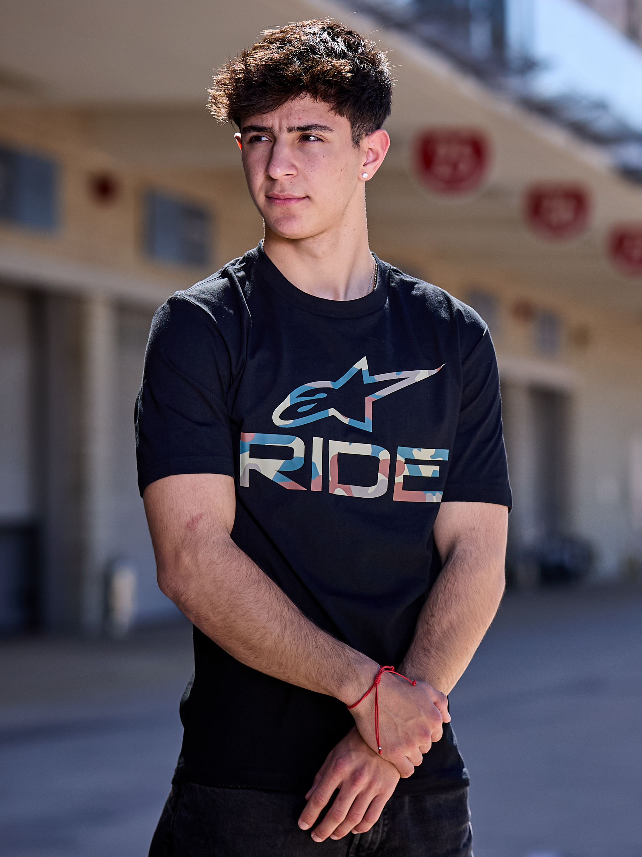 A person stands facing forward, wearing the Alpinestars Ride 4.0 Camo CSF Tee. The black T-shirt features a soft hand print with a colorful graphic logo and the word "RIDE" beneath. The person has short hair and a neutral expression. The background is plain black.