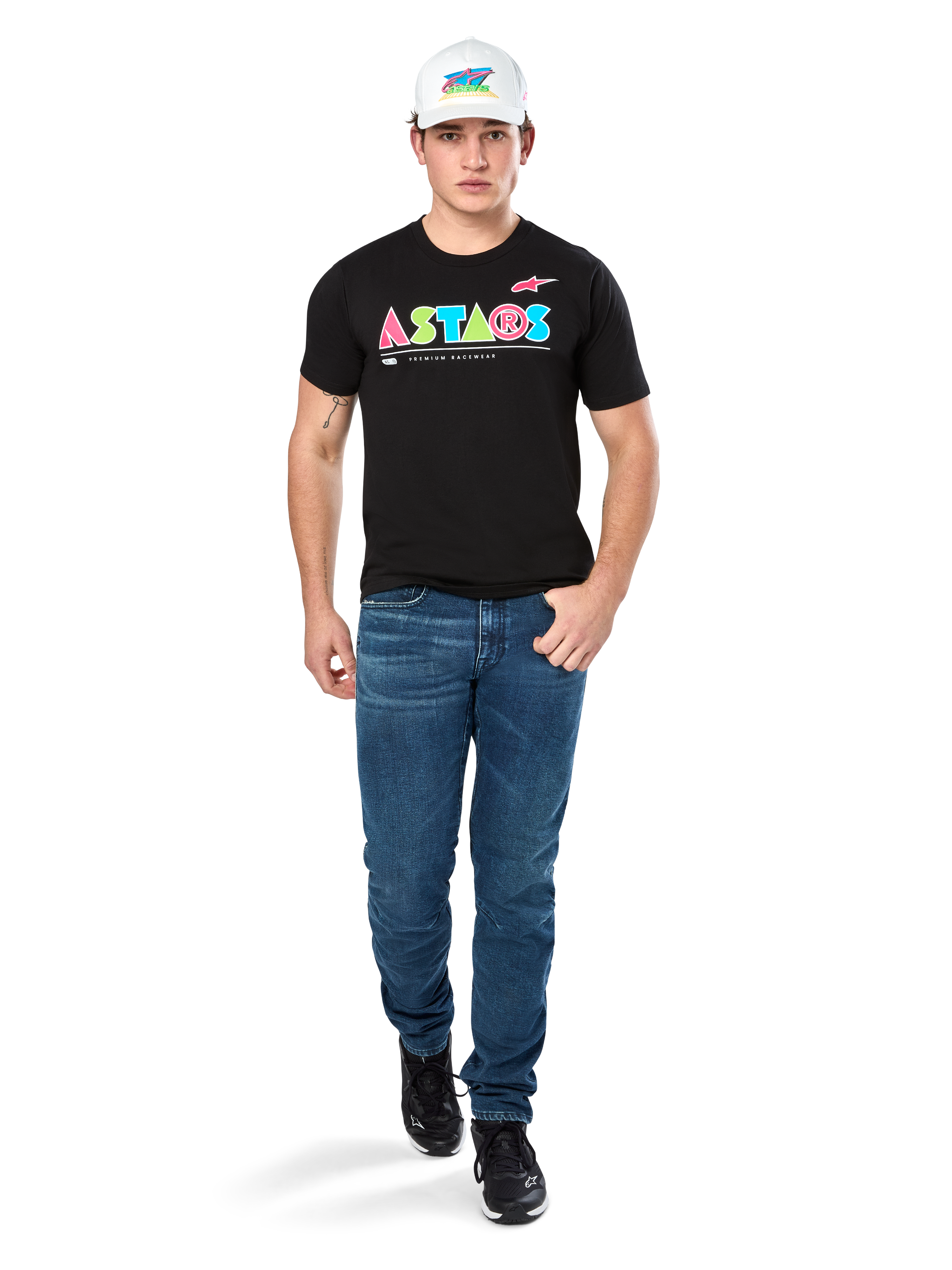 Male model wearing Alpinestars Cereal CSF Tee, Black Short Sleeve T-Shirt with colorful geometric ASTARS logo, paired with blue jeans and white cap, casual motorsport-inspired streetwear.
