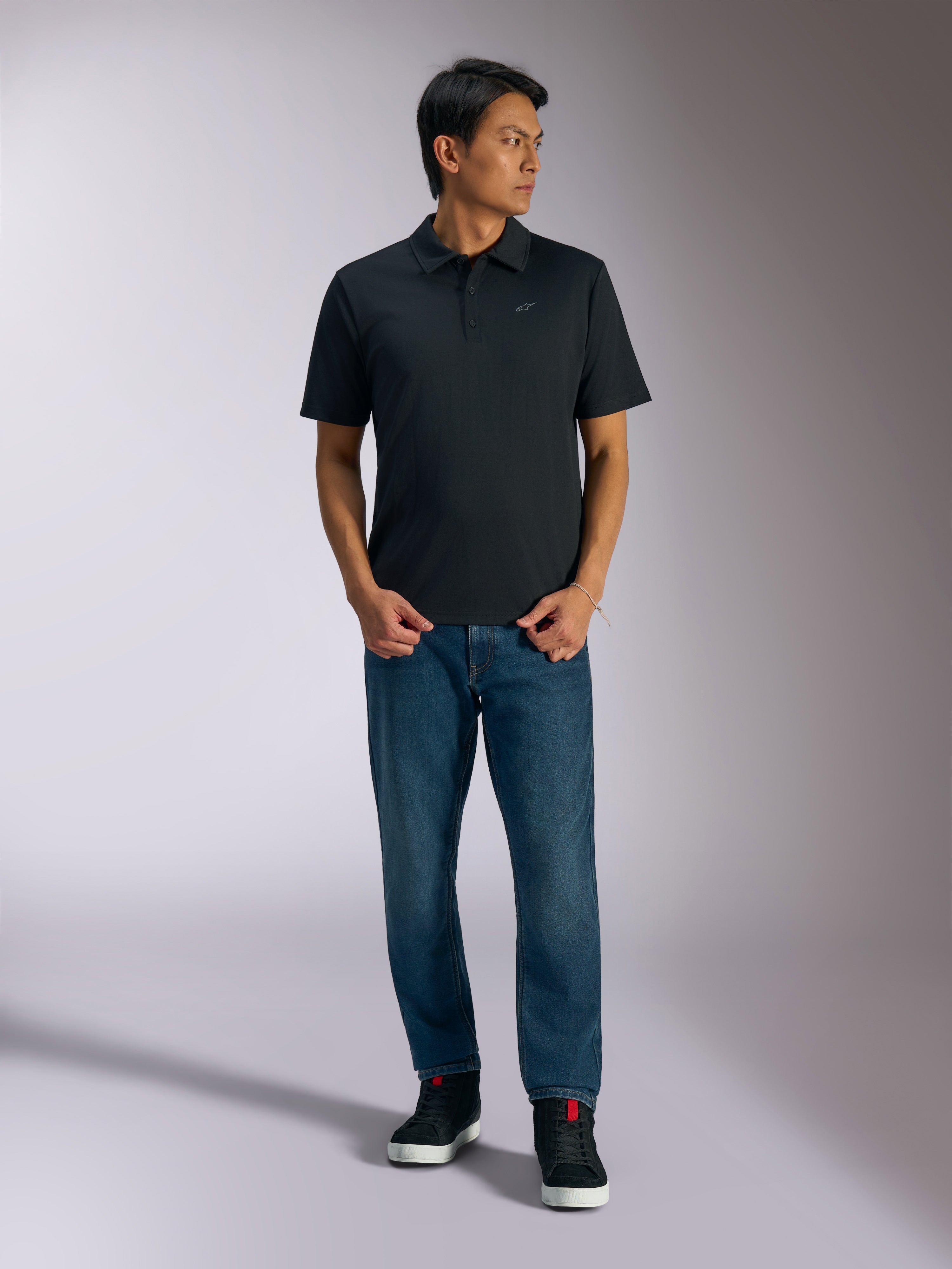 Alpinestars Impetus Sport Polo, Black short sleeve polo shirt, features a three-button placket and grey Astars chest logo, worn by a man with blue jeans and black high-top sneakers, casual motorsport-inspired sportswear