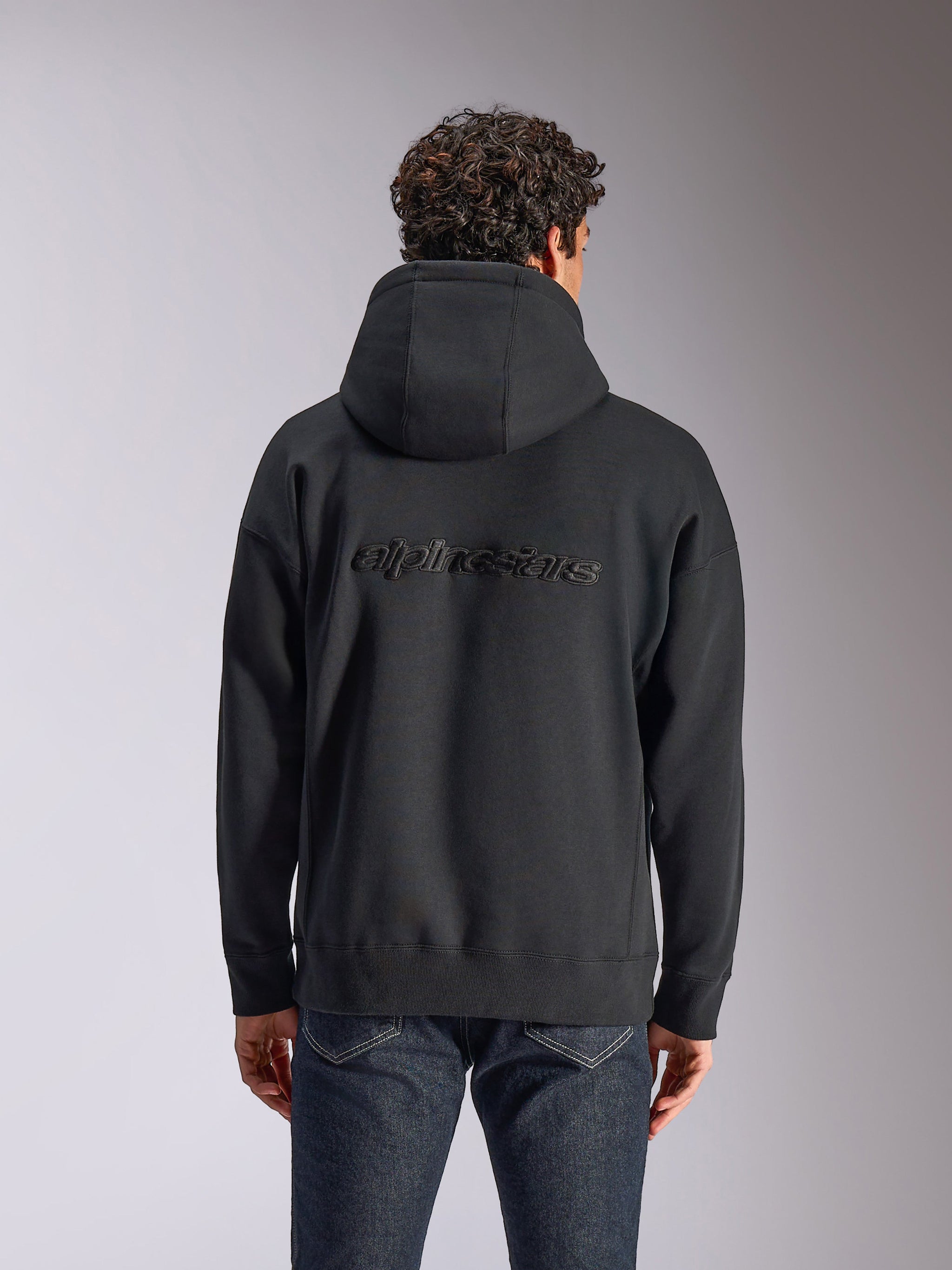 Alpinestars Emergent Oversized Hoodie, back view of a black pullover hoodie on a model, featuring embroidered Alpinestars word logo on the back, oversized fit, casual streetwear paired with dark jeans