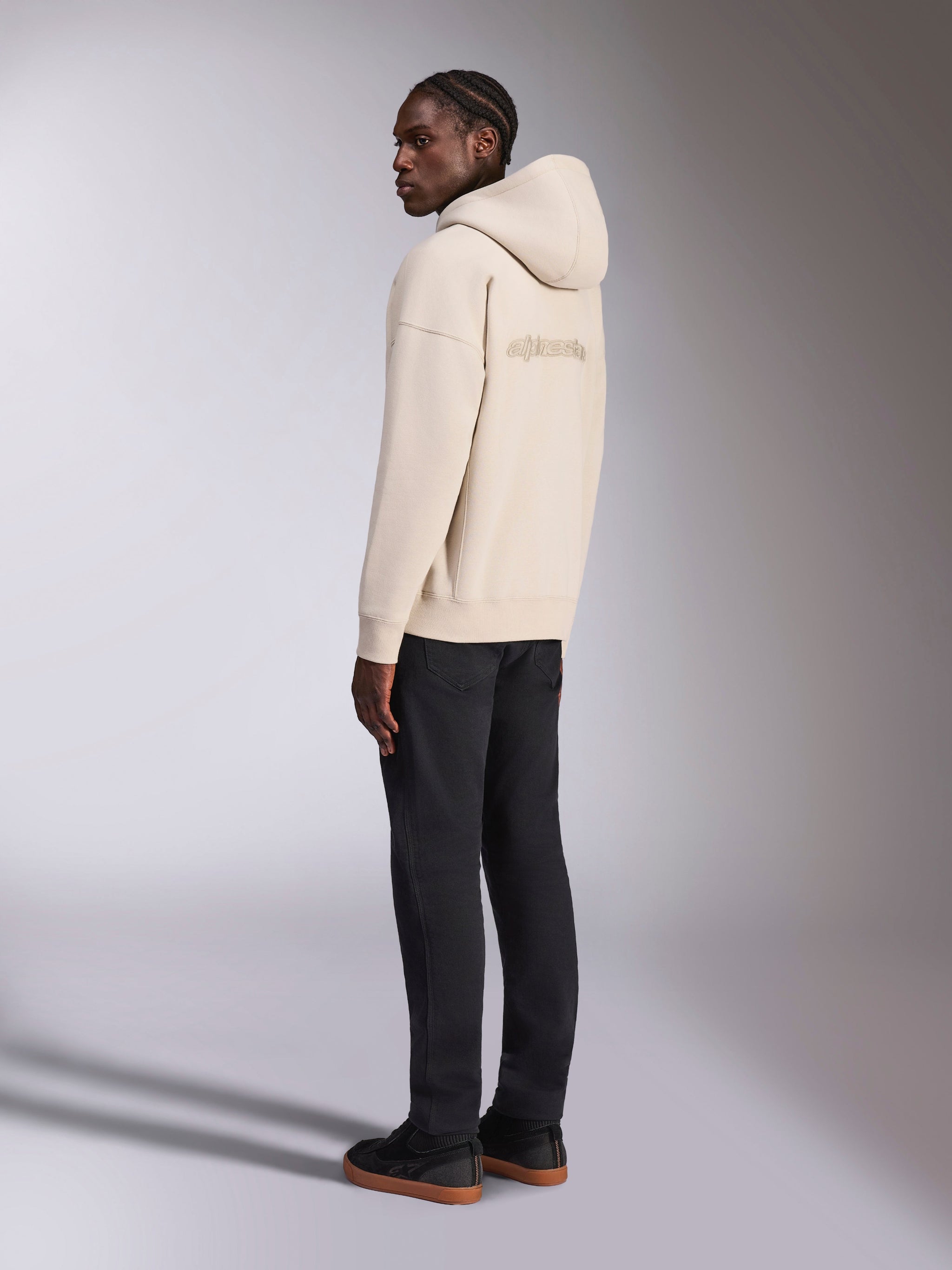 Alpinestars Emergent Oversized Hoodie in Sand, back view showing embroidered Alpinestars logo, oversized fleece pullover with hood, paired with black pants for a casual streetwear look