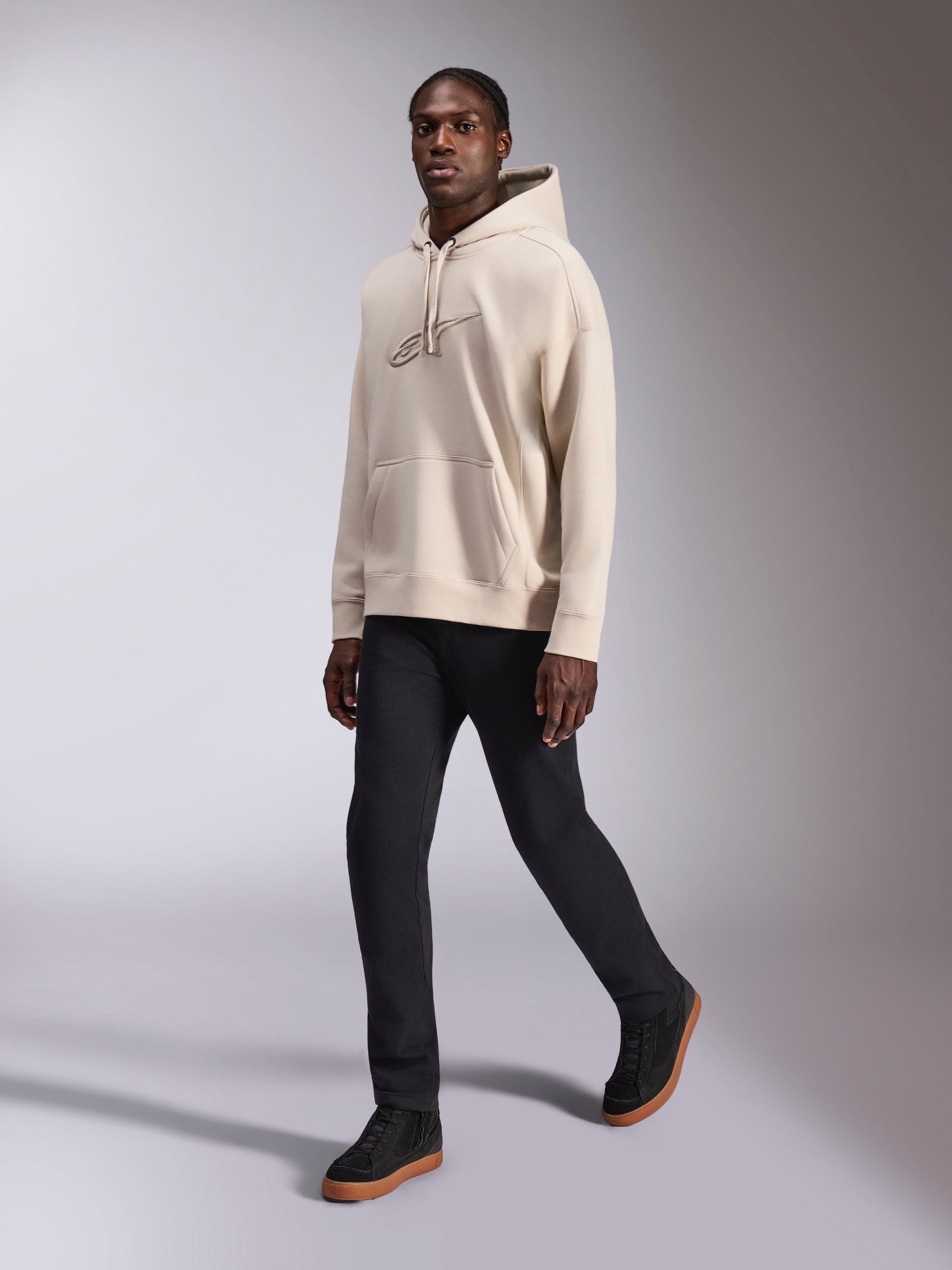 Alpinestars Emergent Oversized Hoodie, casual streetwear, sand off-white, tonal 3D embroidered logo, worn by model with black pants and black sneakers, relaxed oversized fit with kangaroo pocket and drawstring hood