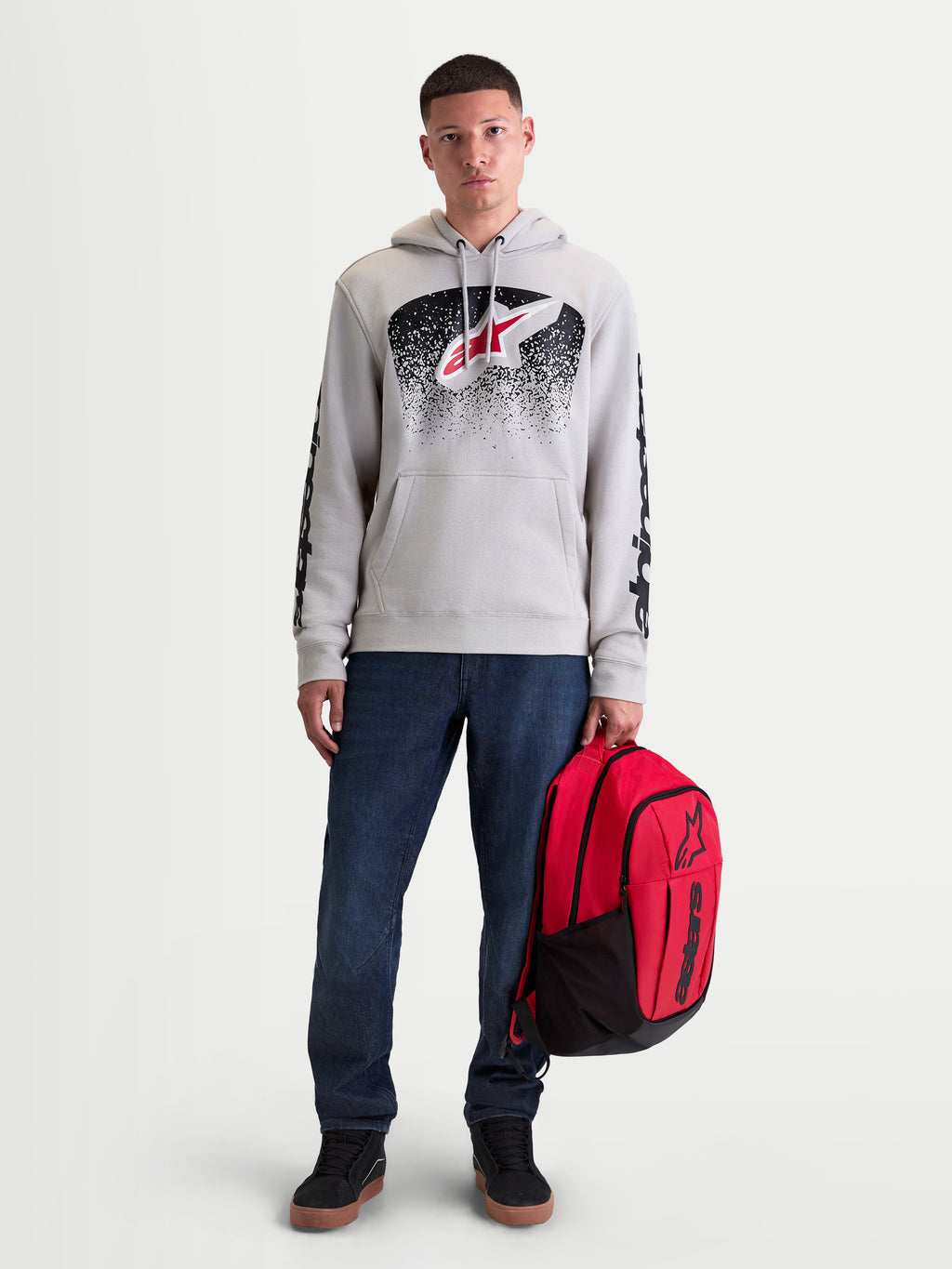 Alpinestars Obsessed Hoodie, Men's Sportswear Pullover Fleece, Cement Gray with Red and Black MX-inspired gradient graphics. This casual motorsport lifestyle hoodie features sleeve branding and is shown on a male model holding a red Alpinestars backpack.