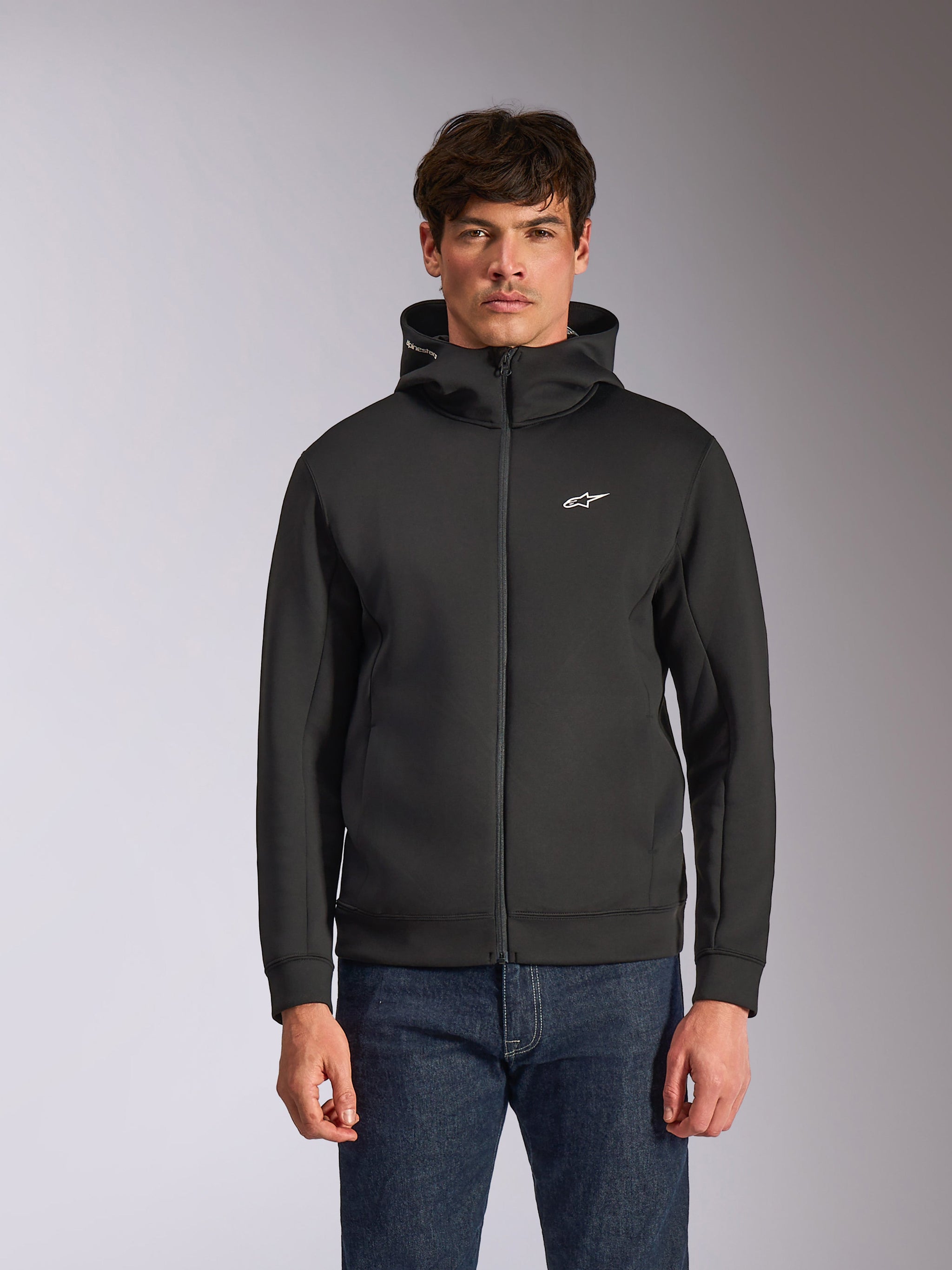 Alpinestars Cypher Hoodie, black zip-front fleece hoodie, featuring a high-neck hood with white branding and a small white Astars logo on the chest, worn by a model with blue jeans, casual motorsport-inspired streetwear.