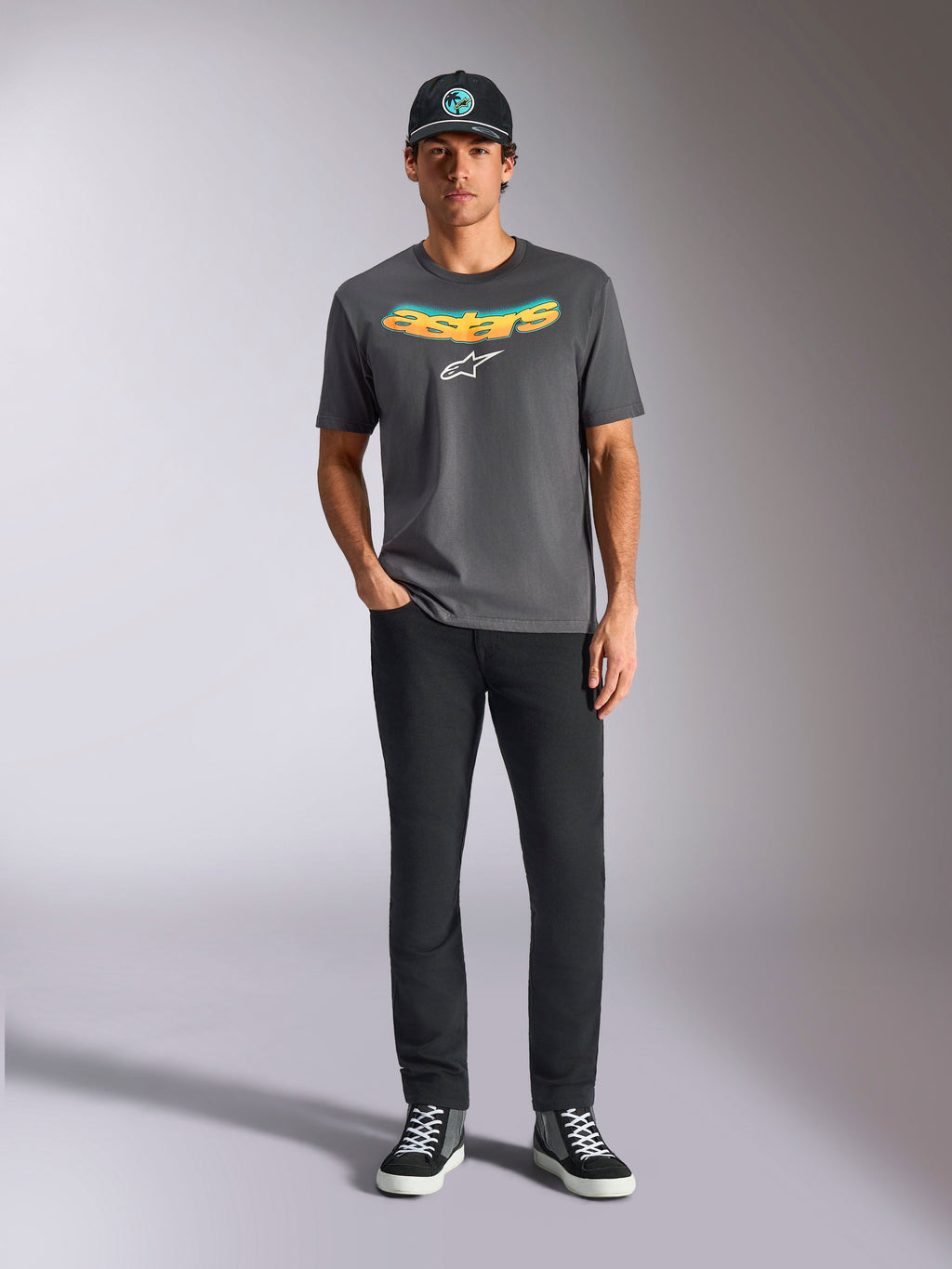 Alpinestars Ellipsoid CSF Tee, Short Sleeve T-Shirt, Charcoal Gray, featuring a curved yellow and orange gradient wordmark and white Astars logo, worn by a model with black pants and a baseball cap, casual sportswear style