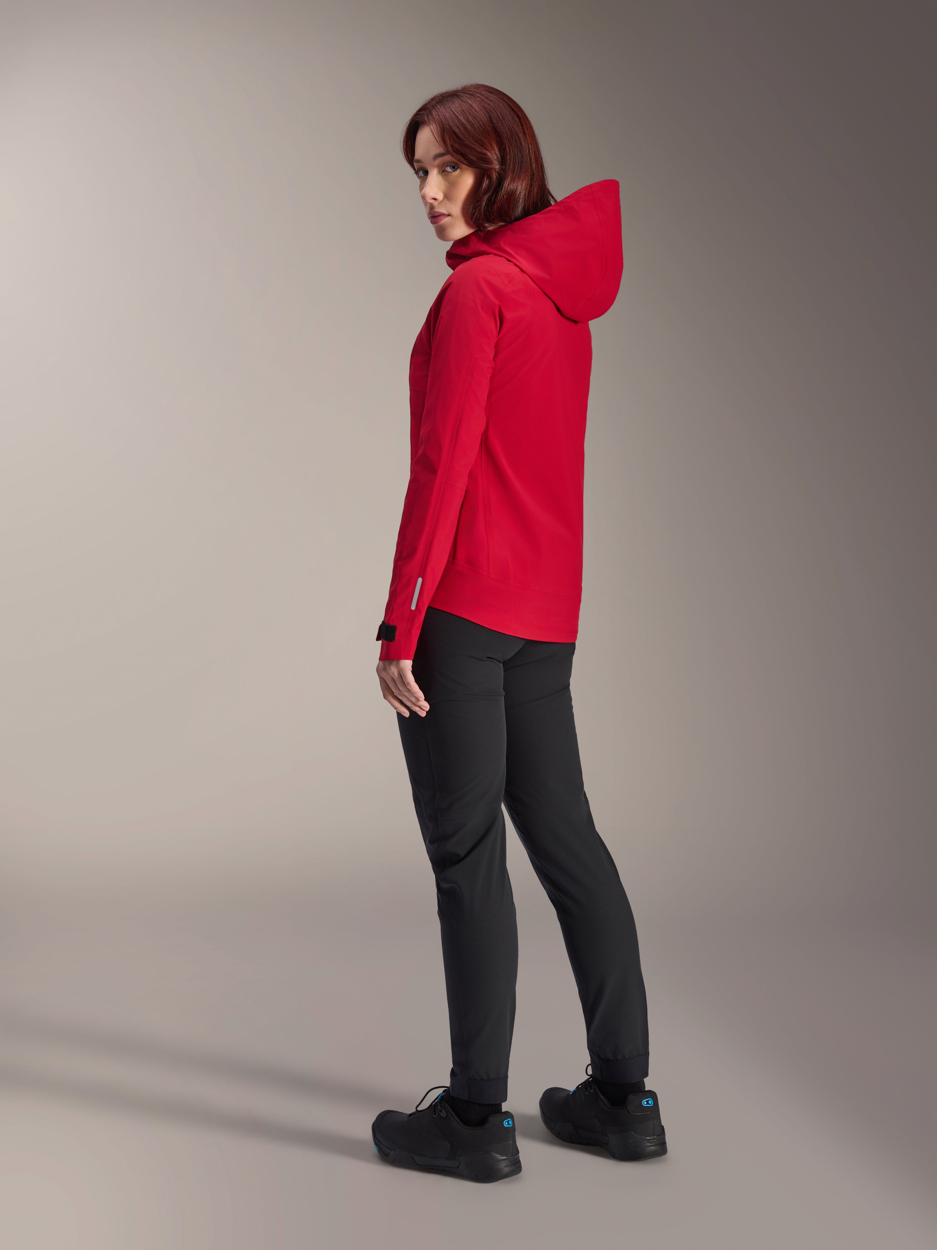 Woman wearing Alpinestars Stella A-Dura Elite Waterproof Jacket in Red Fluo, paired with black technical riding pants and black shoes, rear three-quarter view showing the adjustable hood and dropped tail hem, performance mountain bike apparel.