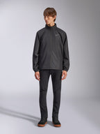 Alpinestars Overhaul Racer Jacket, Black Men's Rain Shell, full-length view on a model paired with black pants and casual sneakers, featuring a small white logo on the chest, water-resistant fabric, and casual motorsport-inspired design