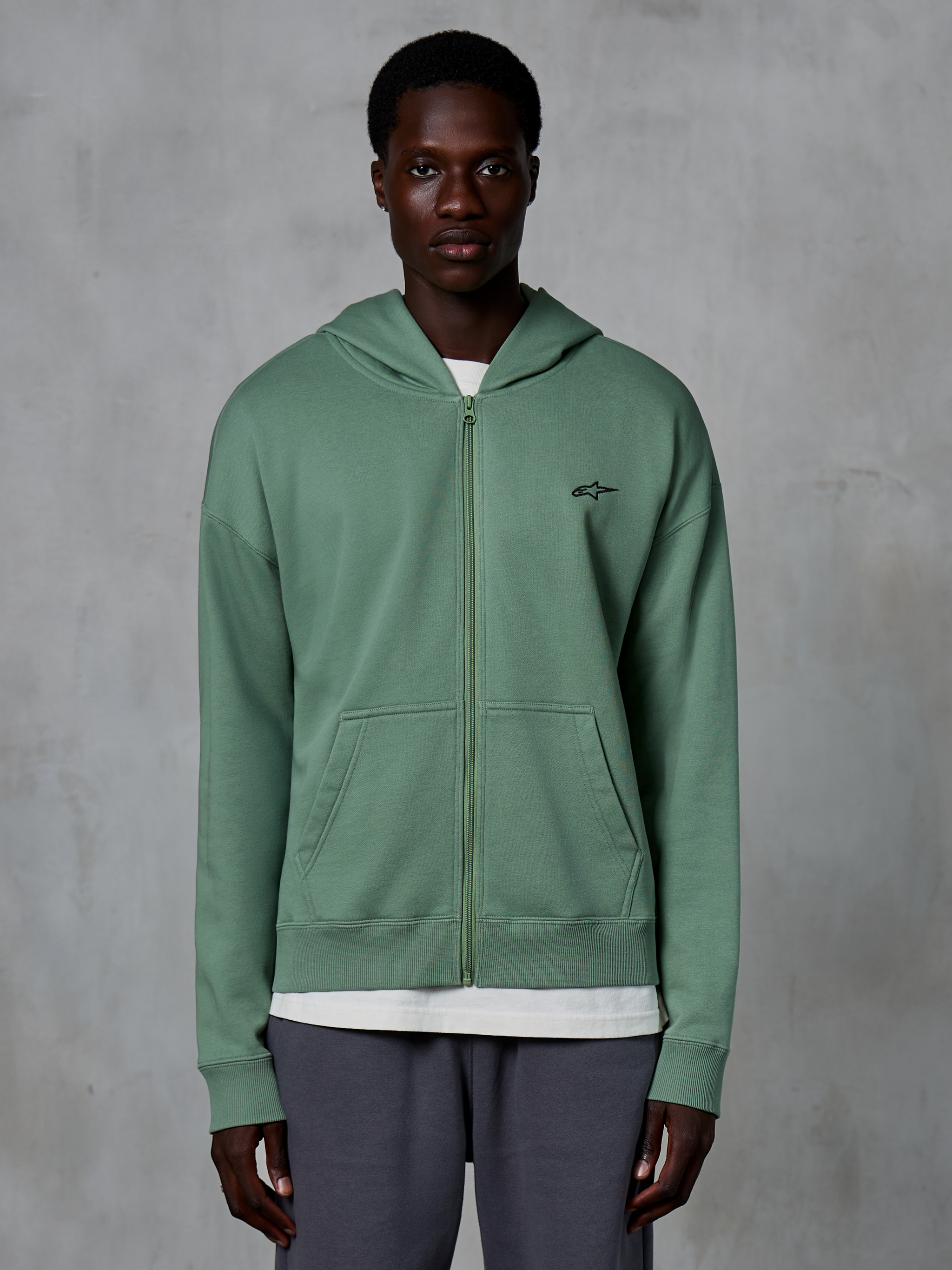 Alpinestars Essentials Ageless Zip Hoodie, casual streetwear zip-up, Cactus green, featuring a subtle embroidered chest logo, split kangaroo pockets, and ribbed cuffs, worn by a male model against a textured grey background