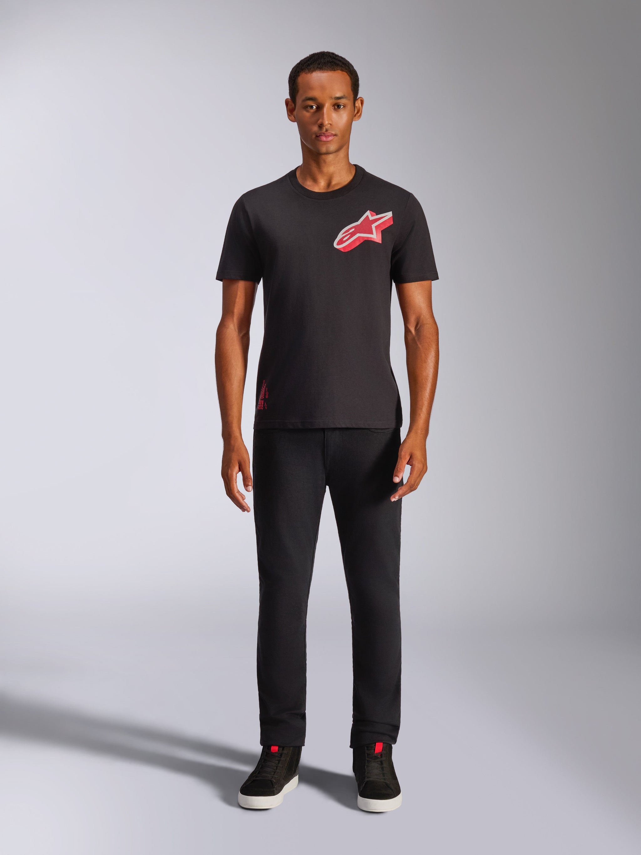 Male model wearing Alpinestars Shaded CSF Tee, Short Sleeve T-Shirt in Black, featuring a red slanted gradient foil Astars logo on the chest, styled with black pants and sneakers, casual motorsport streetwear