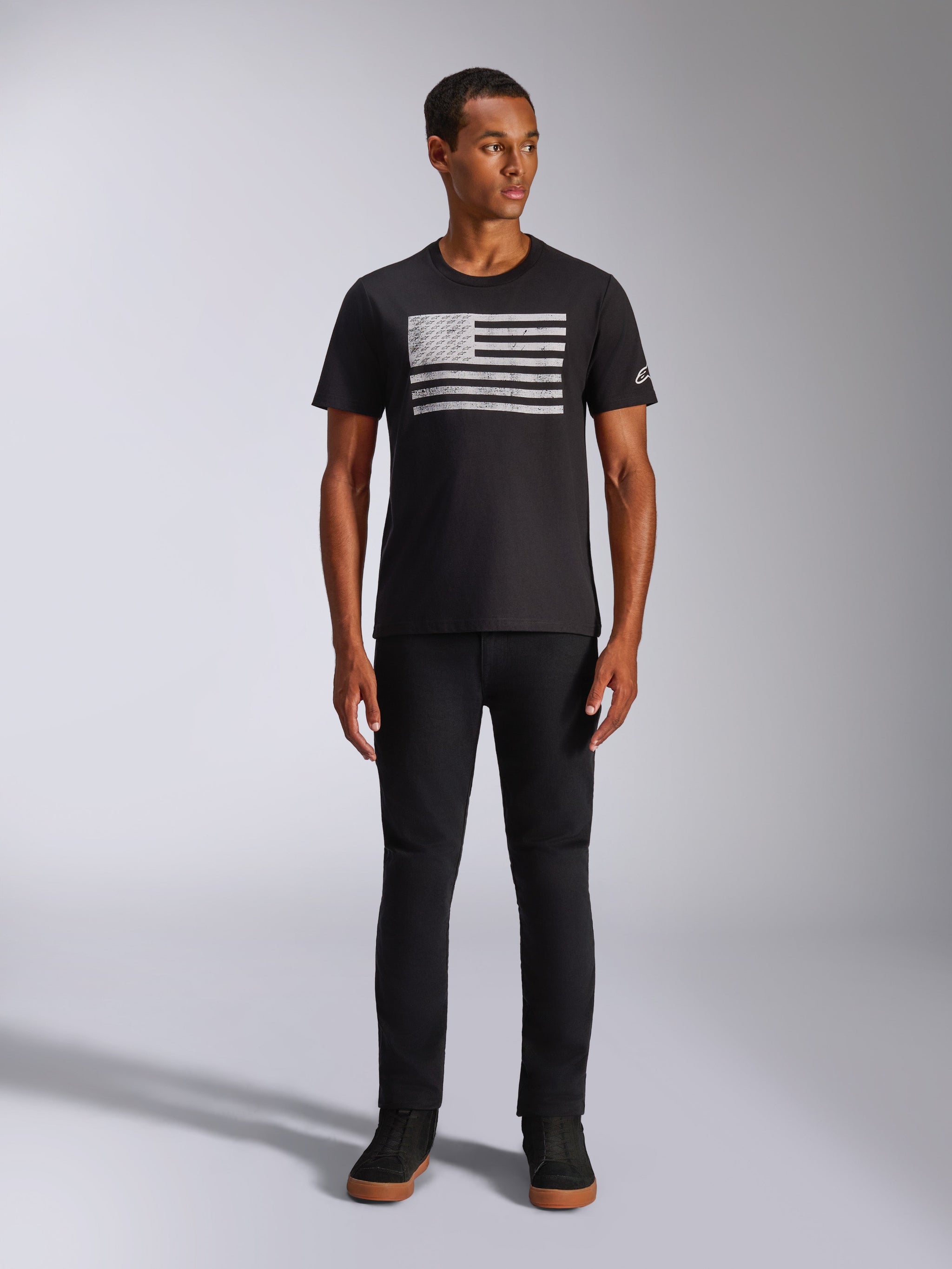 Alpinestars Waving CSF Tee, Short Sleeve T-Shirt, Black, featuring a white distressed flag-style graphic on the chest, worn by a model with black pants and black high-top sneakers, Casual Motorsport Streetwear