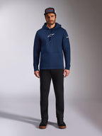 Male model wearing the Alpinestars Frontal Hoodie in Navy, pullover fleece with a white Astars logo on the chest and word logo on the sleeve, featuring a kangaroo pocket and drawstring hood, paired with black pants and a branded cap, casual motorsport streetwear