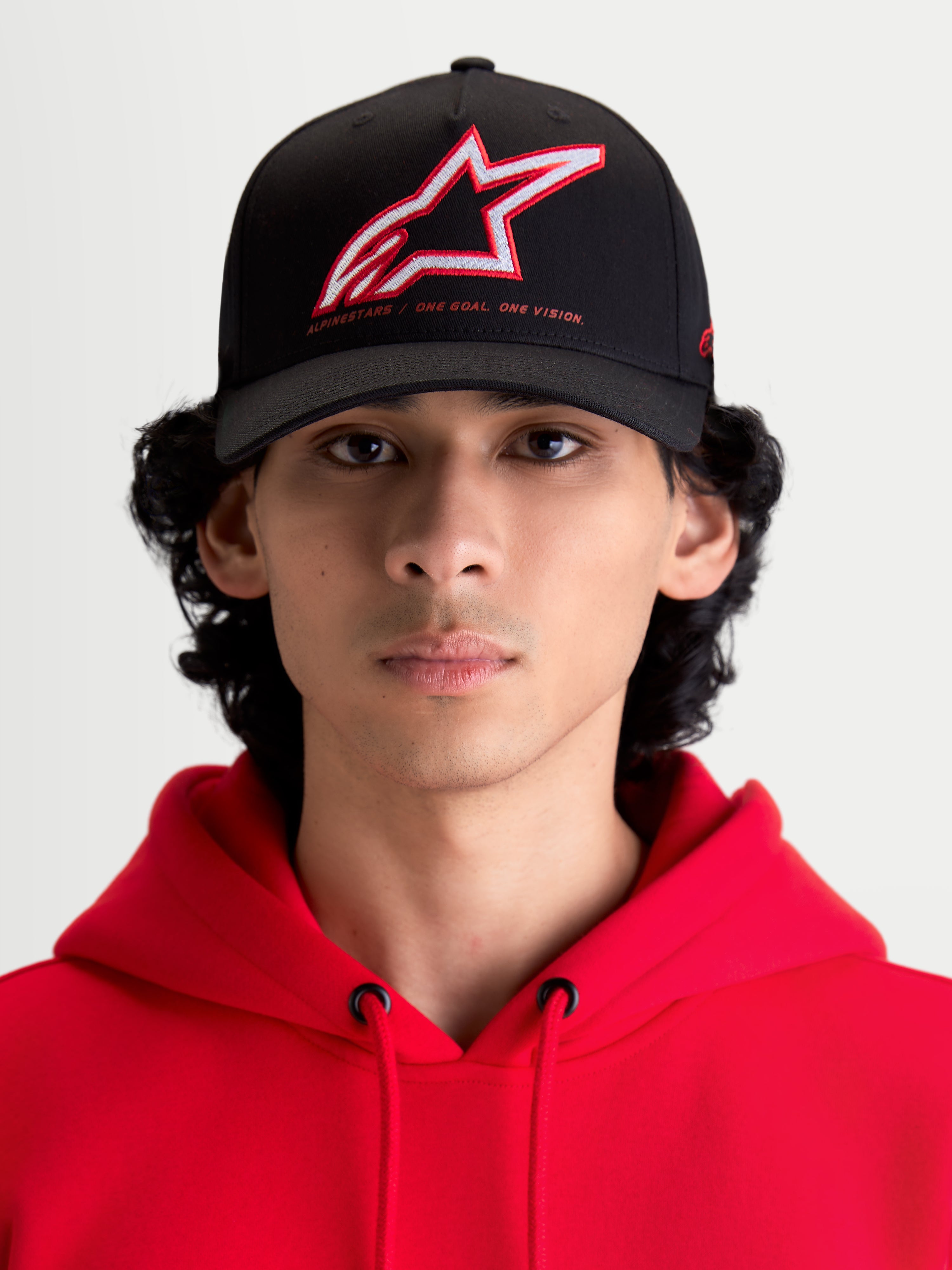 Alpinestars Lineout Hat, Sportswear Curved Bill Baseball Cap, Black with Red and White embroidered Astars logo and One Goal One Vision branding, front view on model wearing a red hoodie, structured twill construction.