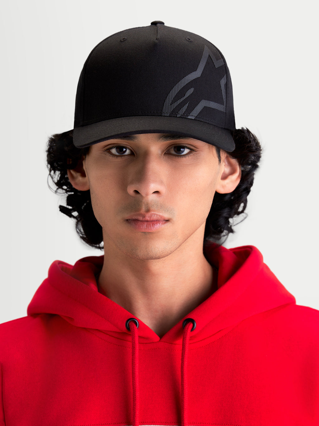 Alpinestars Maze Hat, Black Motorsport-Inspired Sportswear Cap, front view on model wearing a red hoodie. Features a structured crown, curved bill, and a textured Astars Corp Shift logo on durable twill fabric for casual off-bike style.