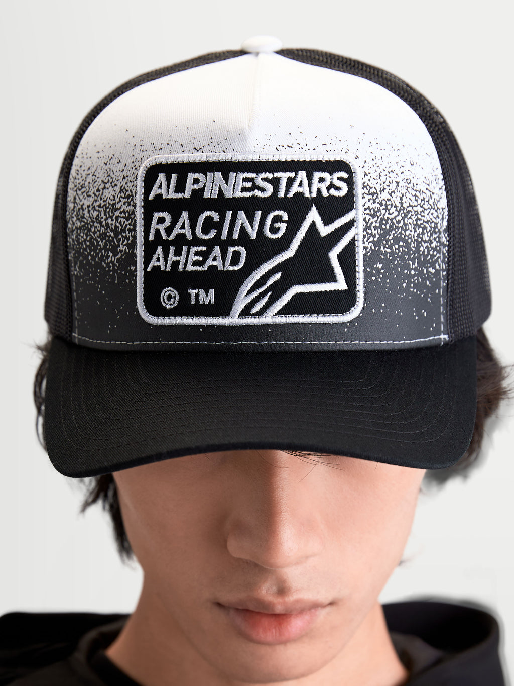 Alpinestars Gravel Trucker Hat, Sportswear Trucker Cap for Men and Women, Black and White, featuring a woven Alpinestars Racing Ahead patch with gravel-texture detailing, structured crown, and curved bill, on-model front view.