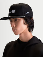 Alpinestars Label Snapback Hat, Black Sportswear Cap with Flat Bill and Structured Crown, featuring a front woven label, white accent rope, and side logo embroidery, shown on model for a modern motorsports-inspired lifestyle look.